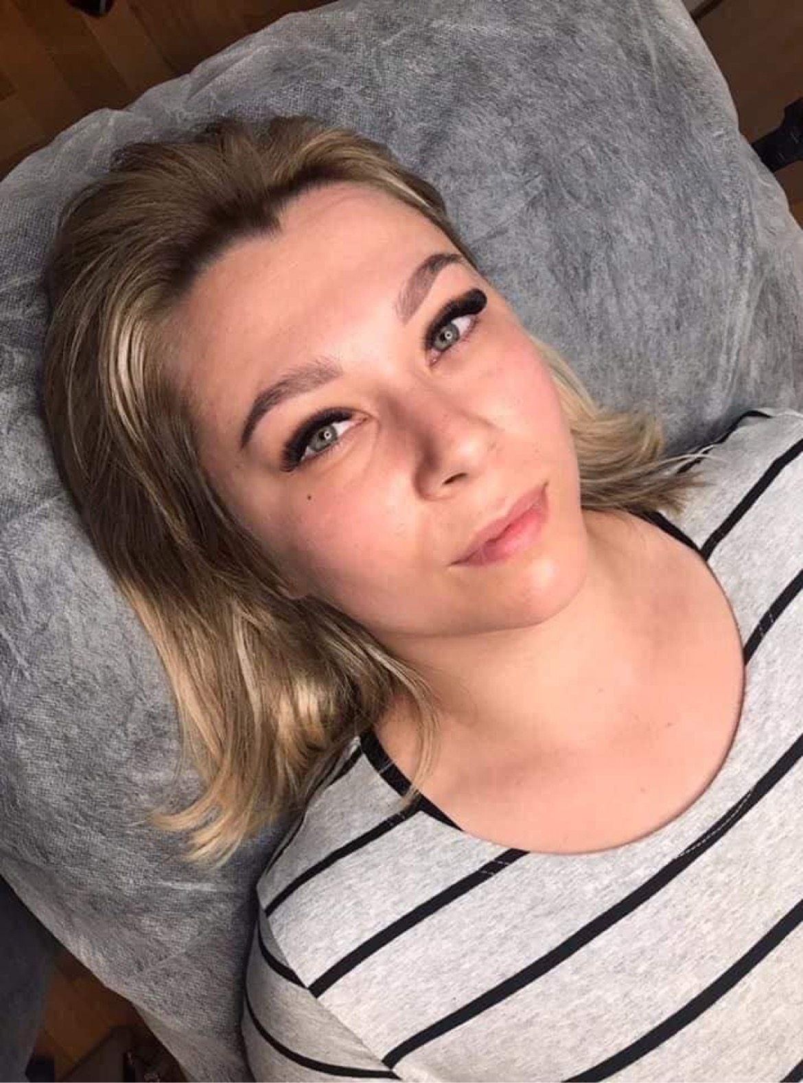 Zbliżenie na twarz kobiety z przedłużonymi rzęsami, leżącej na szarym podłożu. Widoczne podkreślone brwi i delikatny makijaż.