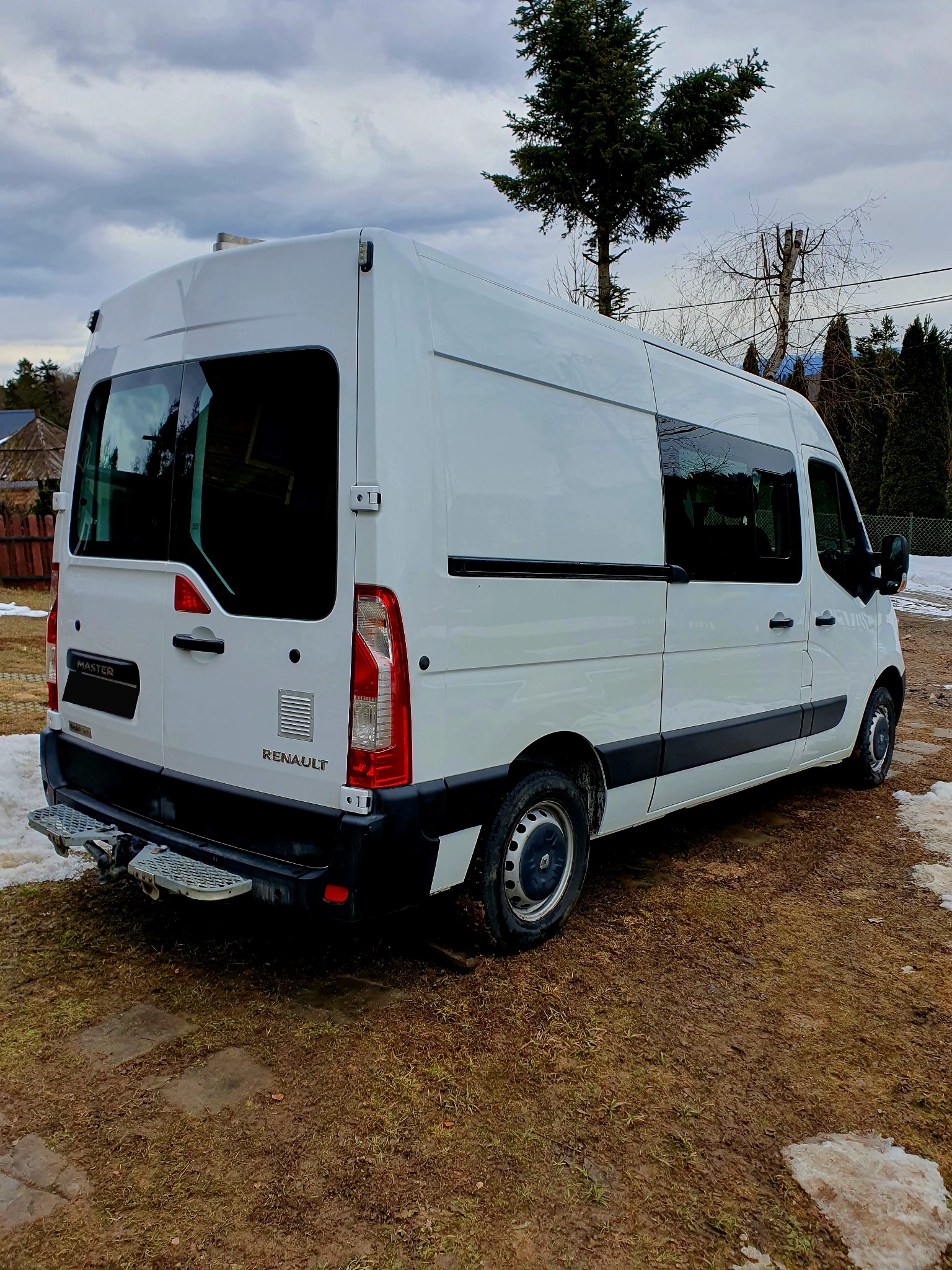 Biały Renault Master z widocznymi stopniami z tyłu, stojący na błotnistym terenie z resztkami śniegu w pochmurny dzień.