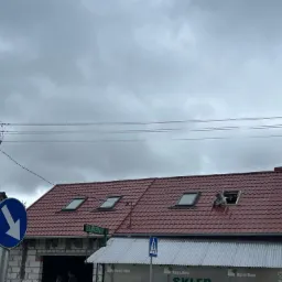 Czerwony dach z oknami dachowymi, z jednym oknem w trakcie montażu przez pracownika, widoczne elementy konstrukcyjne dachu i ciemne chmury na niebie.