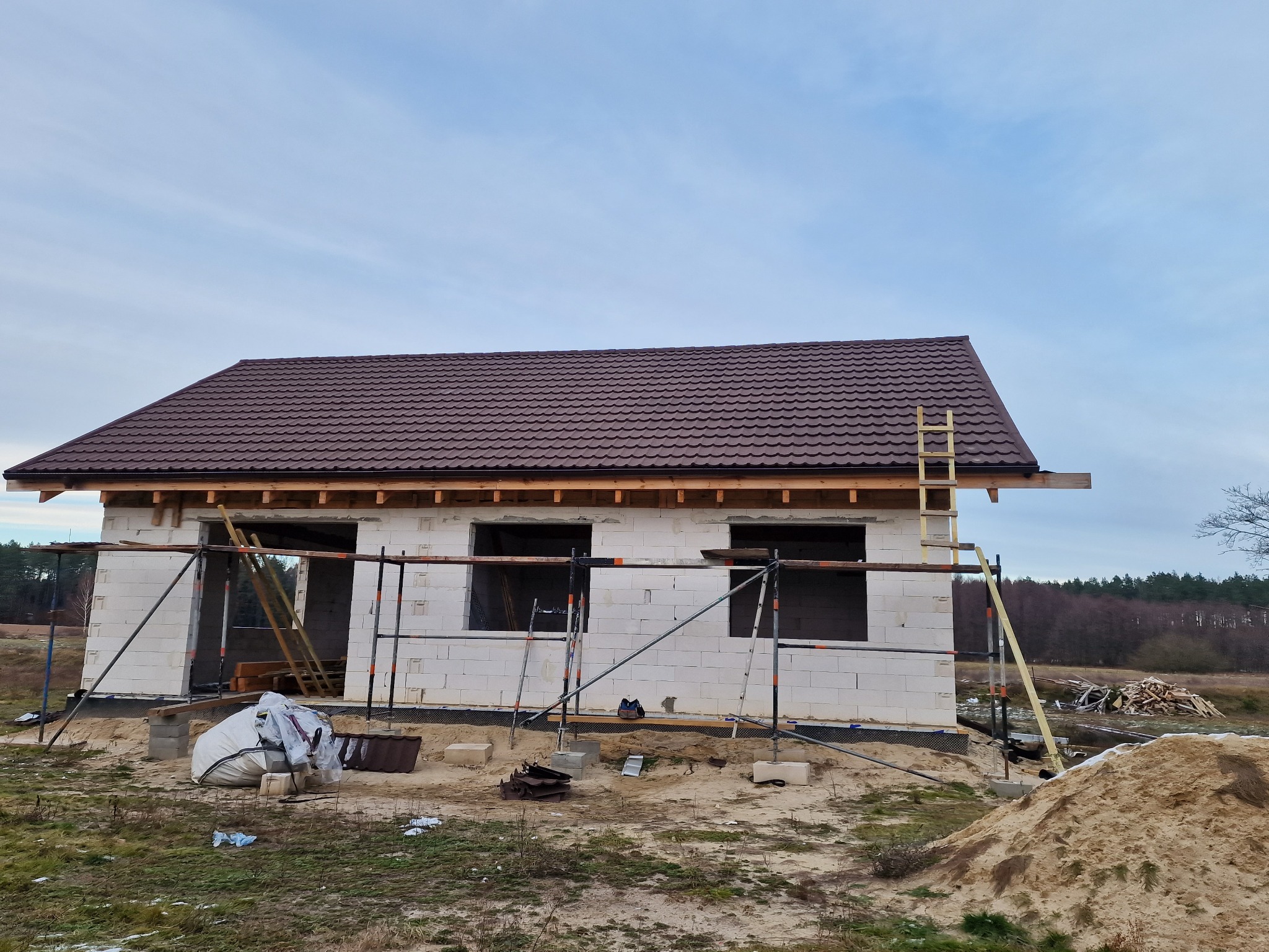 Niedokończony dom jednorodzinny z białych bloczków, z brązowym dachem z blachodachówki, na placu budowy z rusztowaniami i stertą piasku.