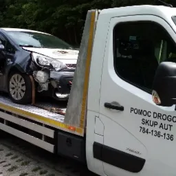 Uszkodzony czarny samochód z naklejką szkoły jazdy transportowany na lawecie pomocy drogowej z widocznym numerem telefonu i napisem 'Skup aut'.