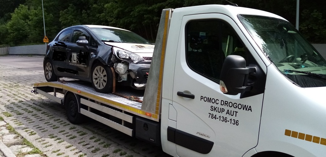 Uszkodzony czarny samochód z naklejką szkoły jazdy transportowany na lawecie pomocy drogowej z widocznym numerem telefonu i napisem 'Skup aut'.