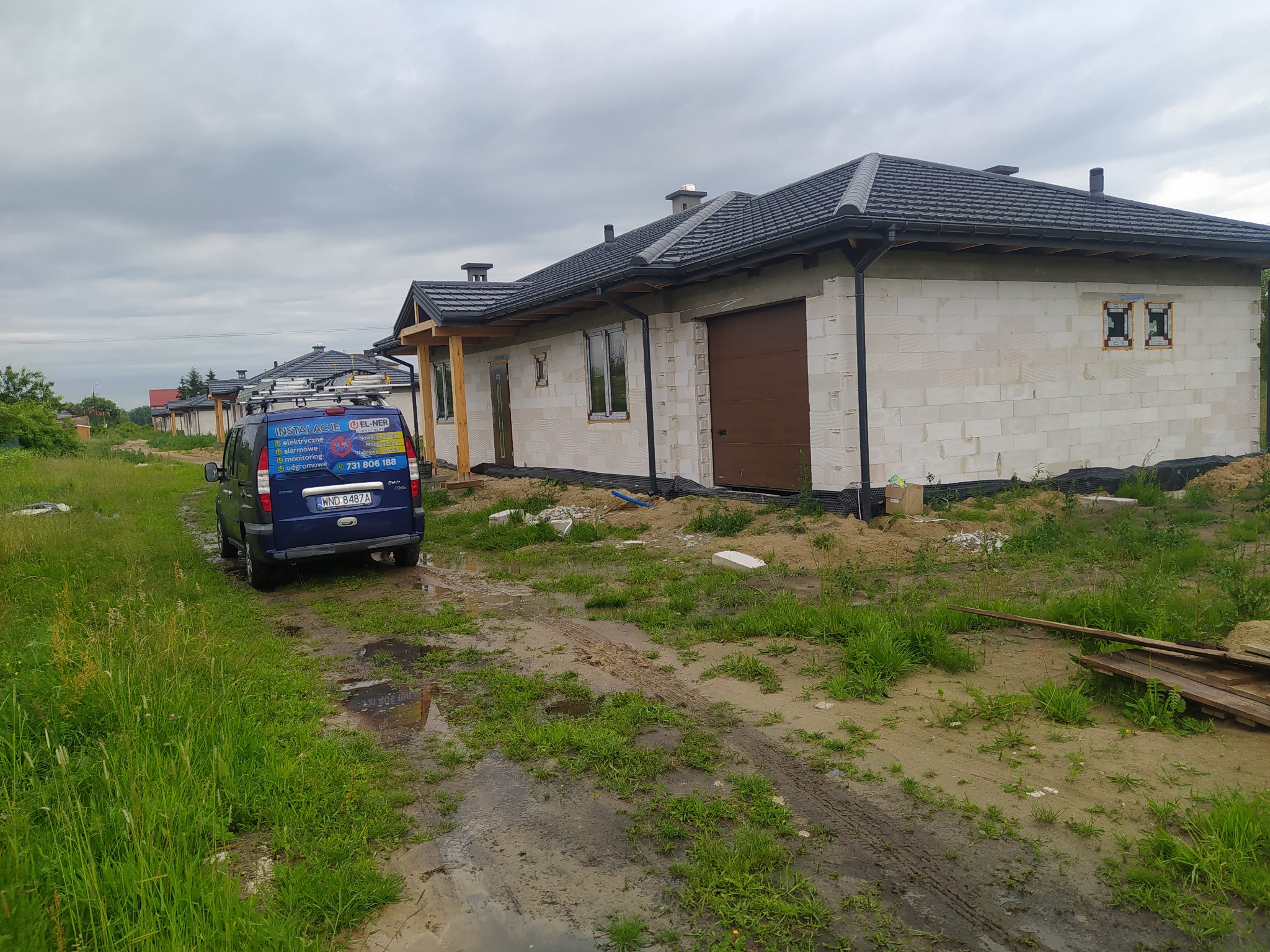 Niebieski samochód firmy instalacyjnej z drabiną na dachu zaparkowany przed szeregiem domów w stanie surowym, widoczne reklamy usług elektrycznych, alarmowych i odgromowych na pojeździe.