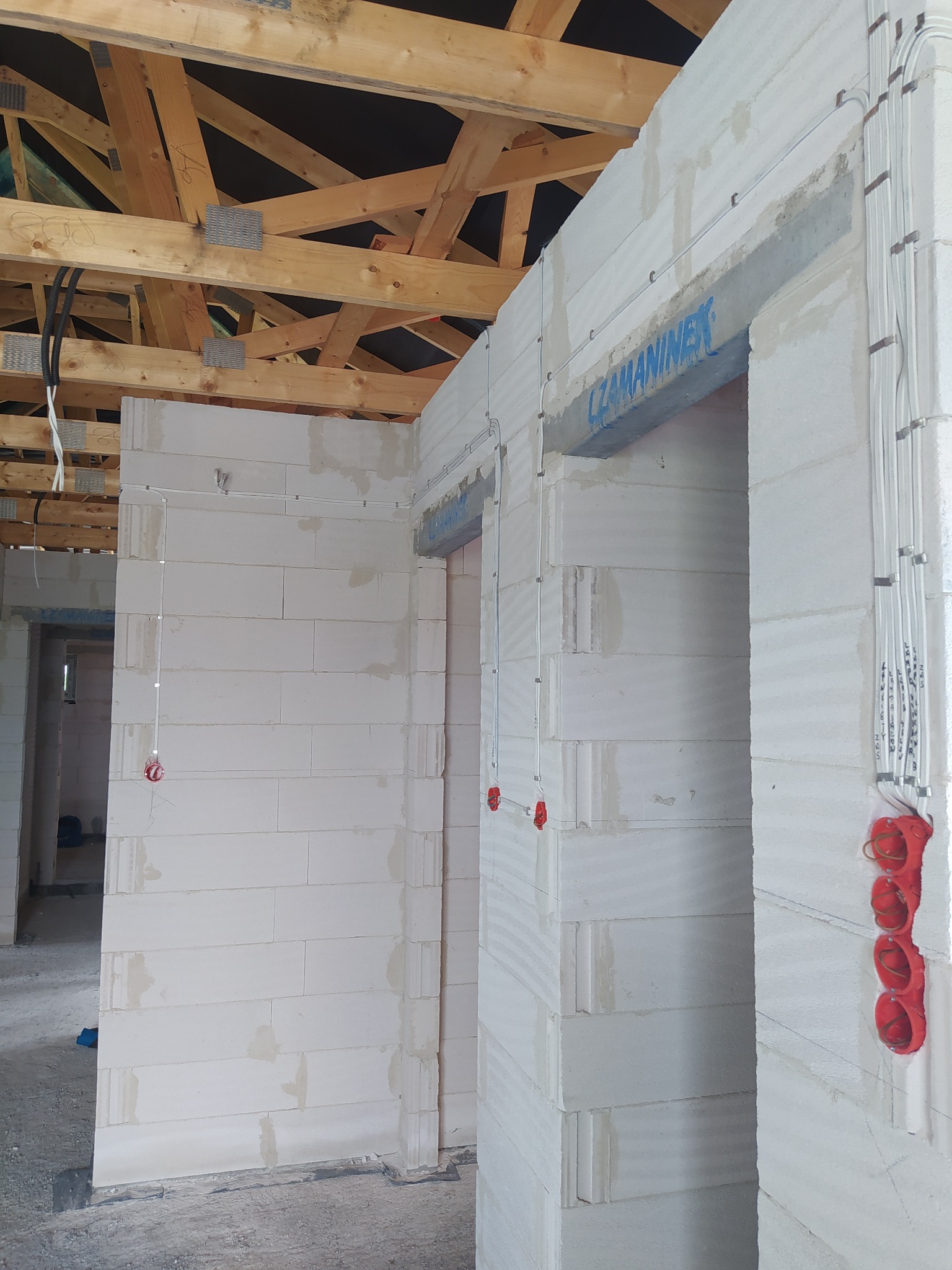 Wewnętrzne ściany budynku w trakcie budowy z widoczną instalacją elektryczną w peszlach i puszkach, drewniana konstrukcja dachu na górze.
