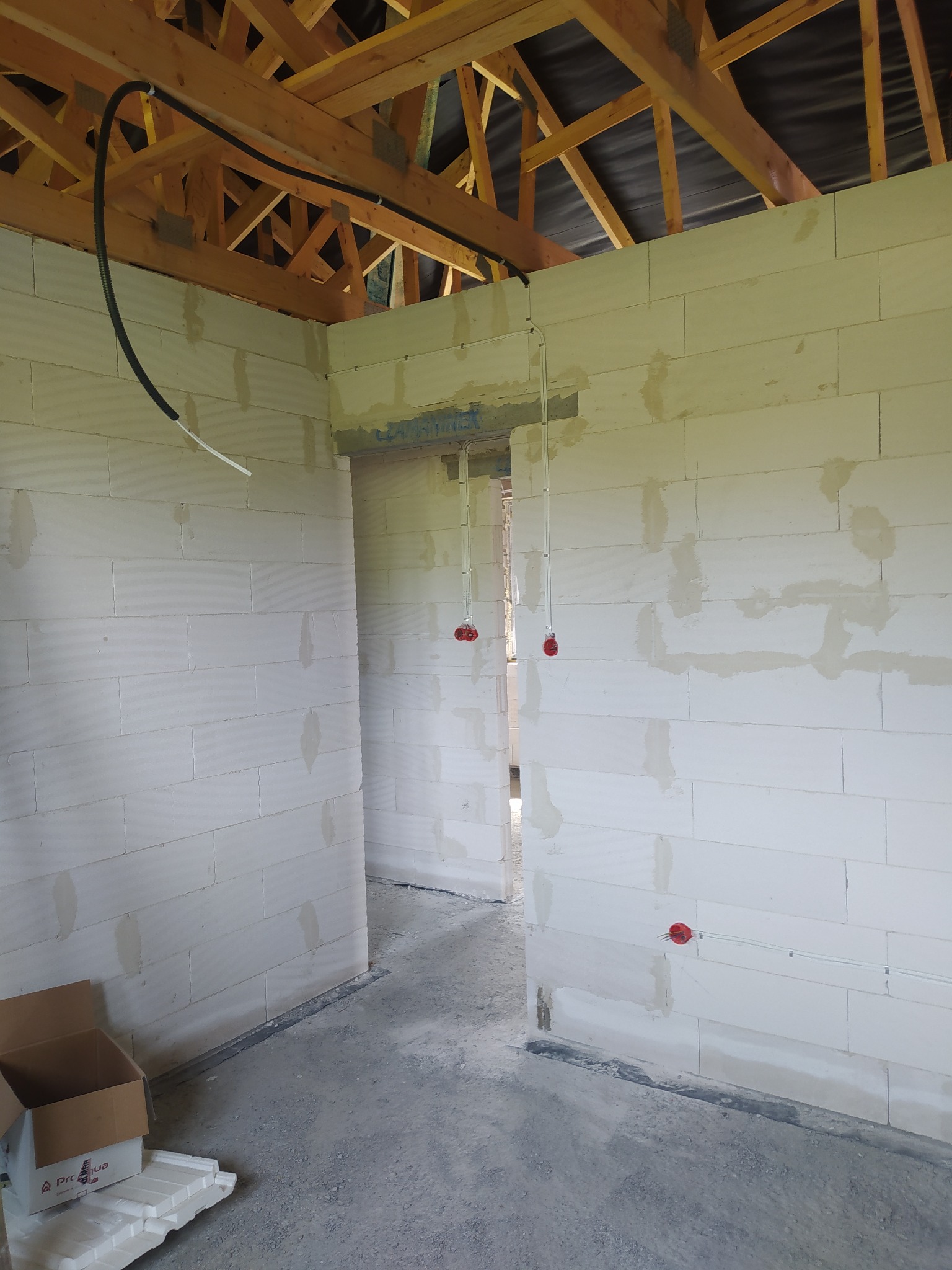 Wnętrze surowego budynku z widoczną instalacją elektryczną: kable prowadzone pod stropem i wypusty na ścianach z czerwonymi puszkami, ściany z bloczków betonowych, surowy strop drewniany.