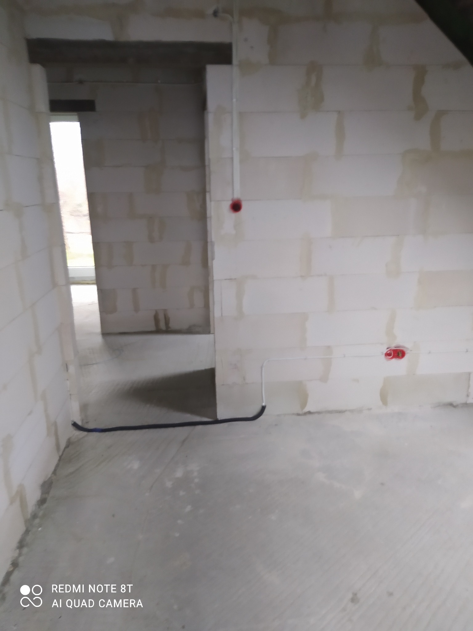 Surowy stan wnętrza z instalacją elektryczną: przewody w peszlu i puszki podtynkowe w ścianie z białych bloczków, widok na sąsiednie pomieszczenie.