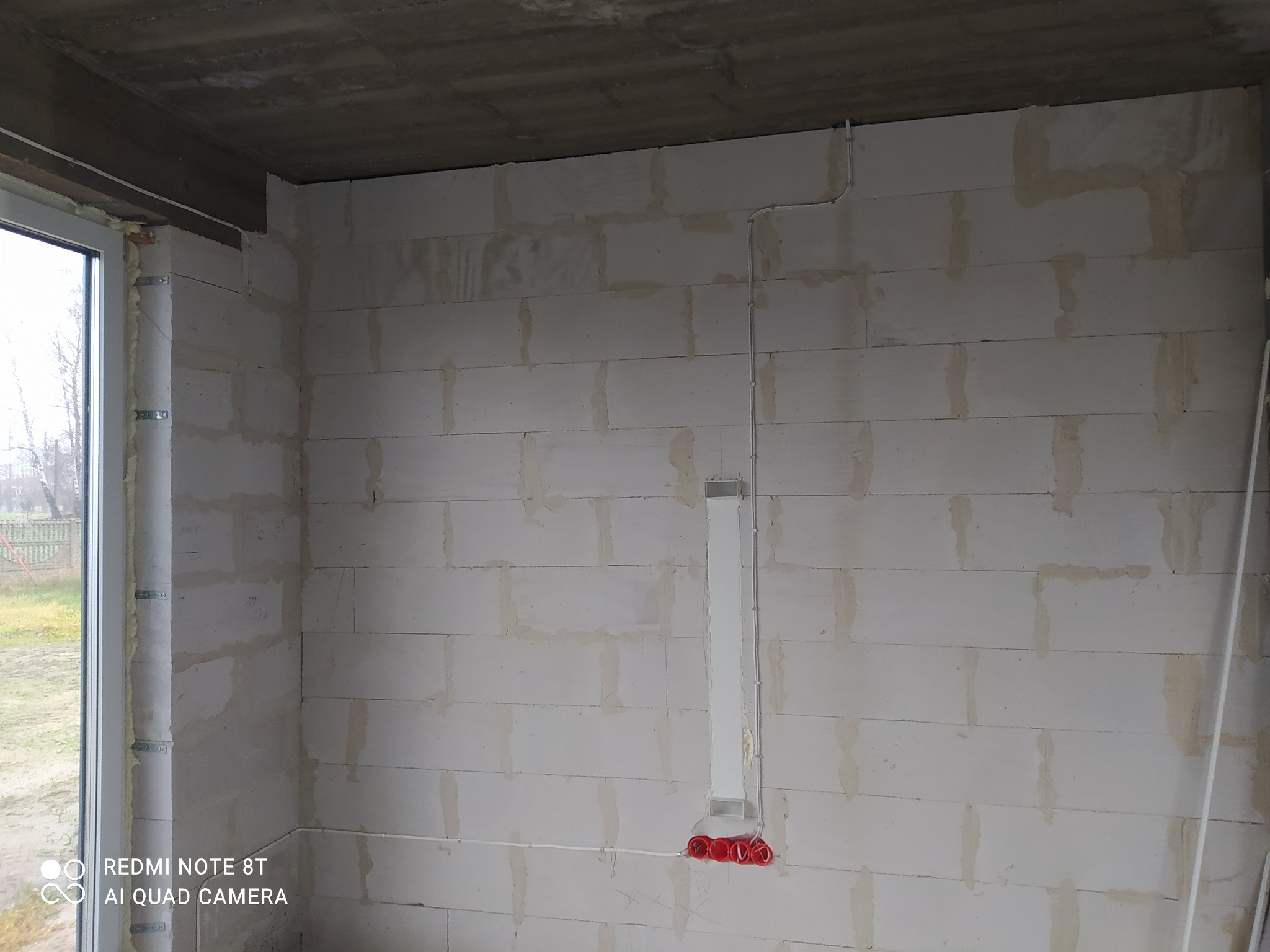 Surowy stan wnętrza w trakcie prac elektrycznych: widoczne przewody elektryczne prowadzone po ścianie z betonu komórkowego, puszki instalacyjne w kolorze czerwonym oraz niezabezpieczone okno...