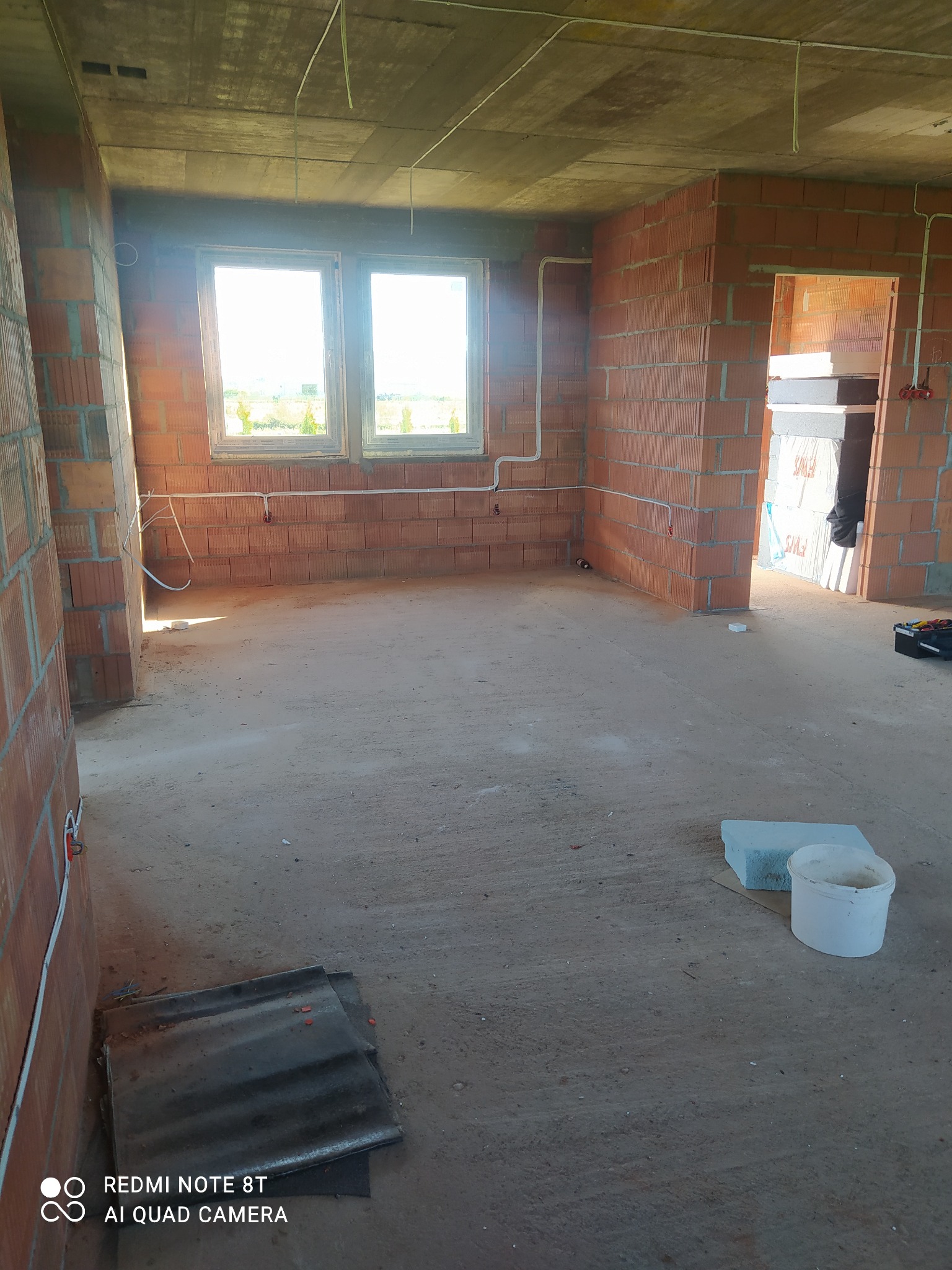 Wnętrze surowego budynku w trakcie prac instalacyjnych: widoczne okablowanie elektryczne poprowadzone po ścianach z czerwonej cegły dziurawki, betonowy sufit, podłoga wylewka, ślady prac...