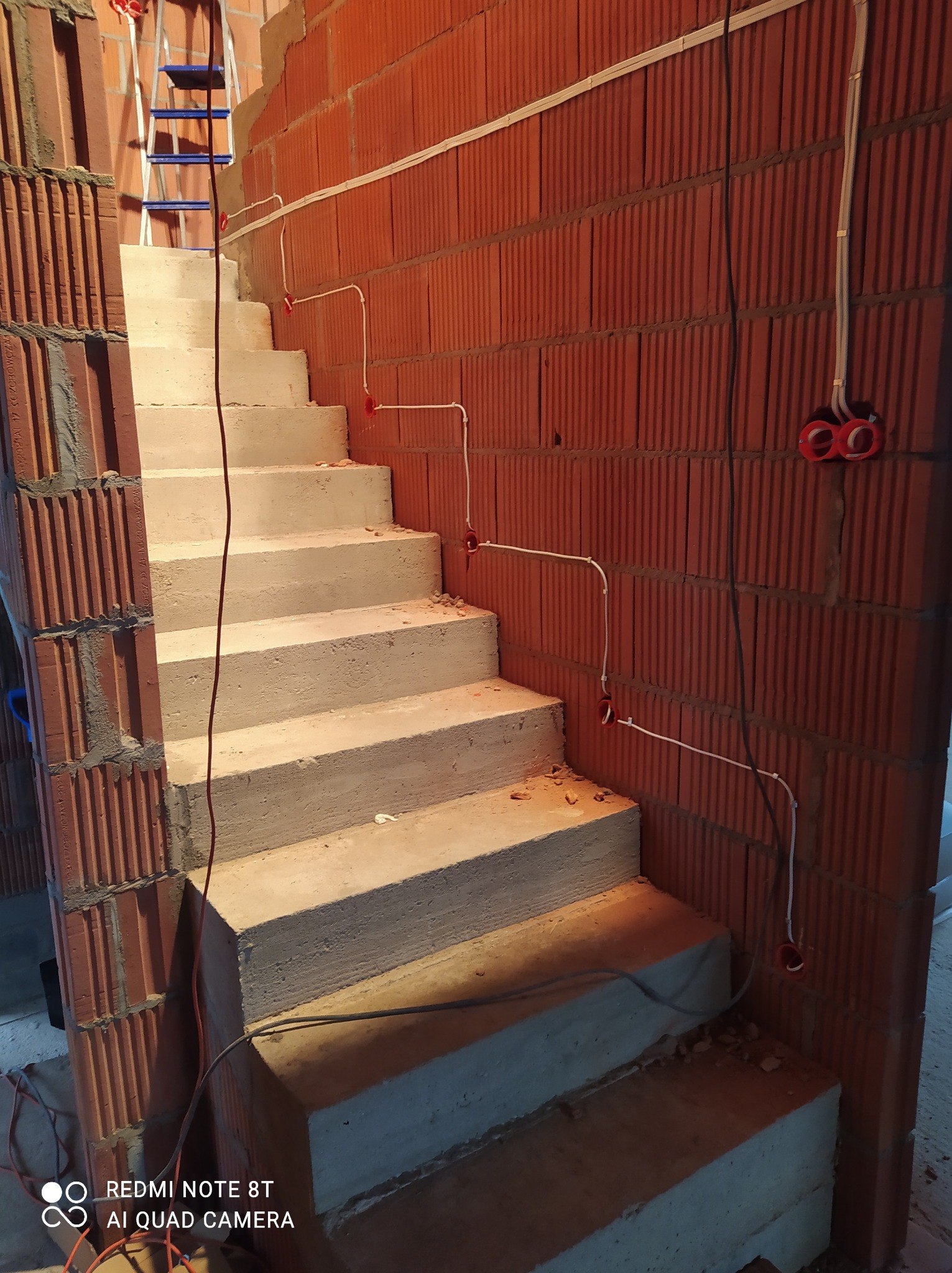 Surowe schody betonowe w trakcie budowy, z widocznymi przewodami elektrycznymi w czerwonych puszkach na ceglanej ścianie i drabiną w tle.