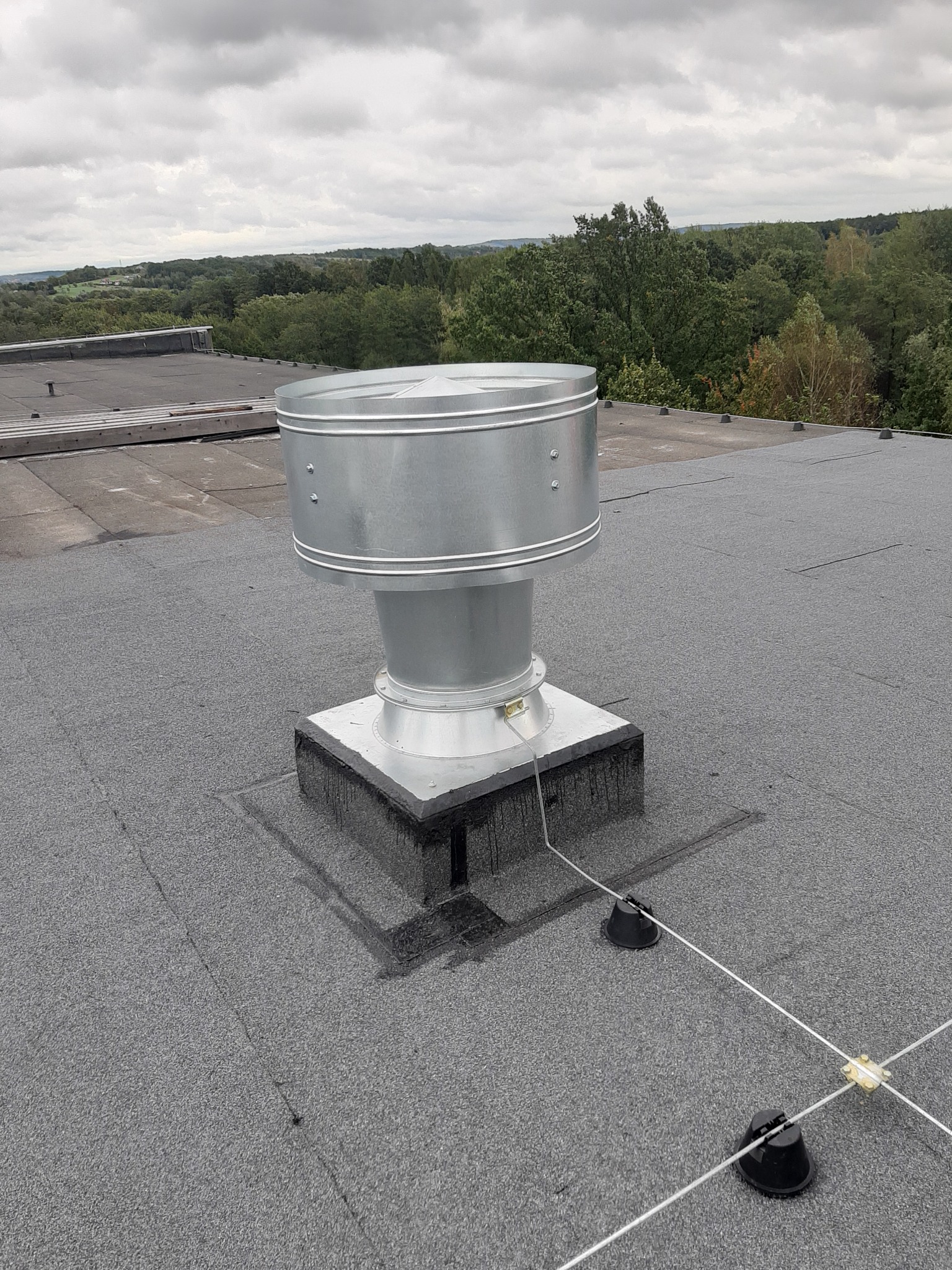 Metalowy komin wentylacyjny na płaskim dachu pokrytym papą z widocznym systemem odgromowym i fragmentem krajobrazu w tle.