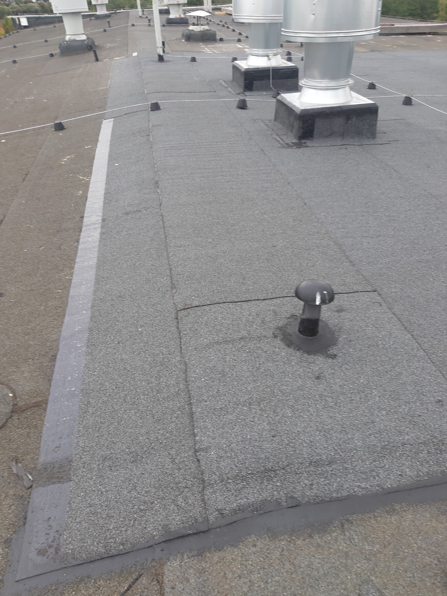 Widok z góry na płaski dach pokryty szarą papą z widocznymi kominkami wentylacyjnymi i zabezpieczeniami odgromowymi.