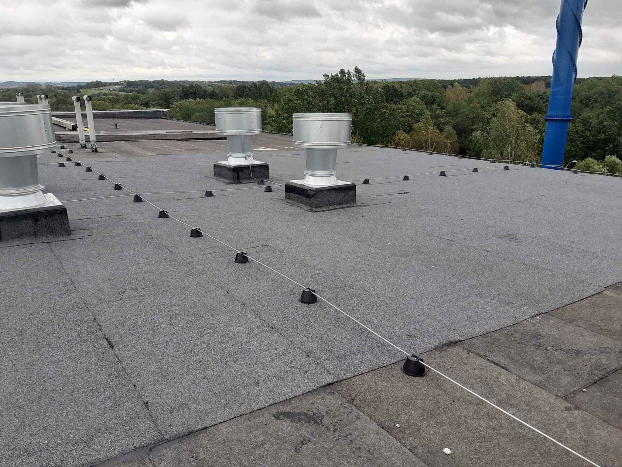 Szary dach hali przemysłowej z wentylacją kominkową zabezpieczony linką asekuracyjną, w tle widok na zalesione tereny pod pochmurnym niebem.