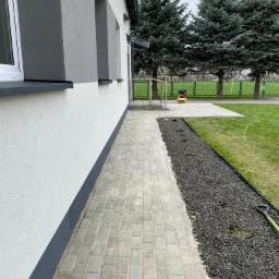 Szary chodnik z kostki brukowej przylegający do ściany budynku, obok rabata z czarnym grysem, w tle trawnik i drzewa iglaste.