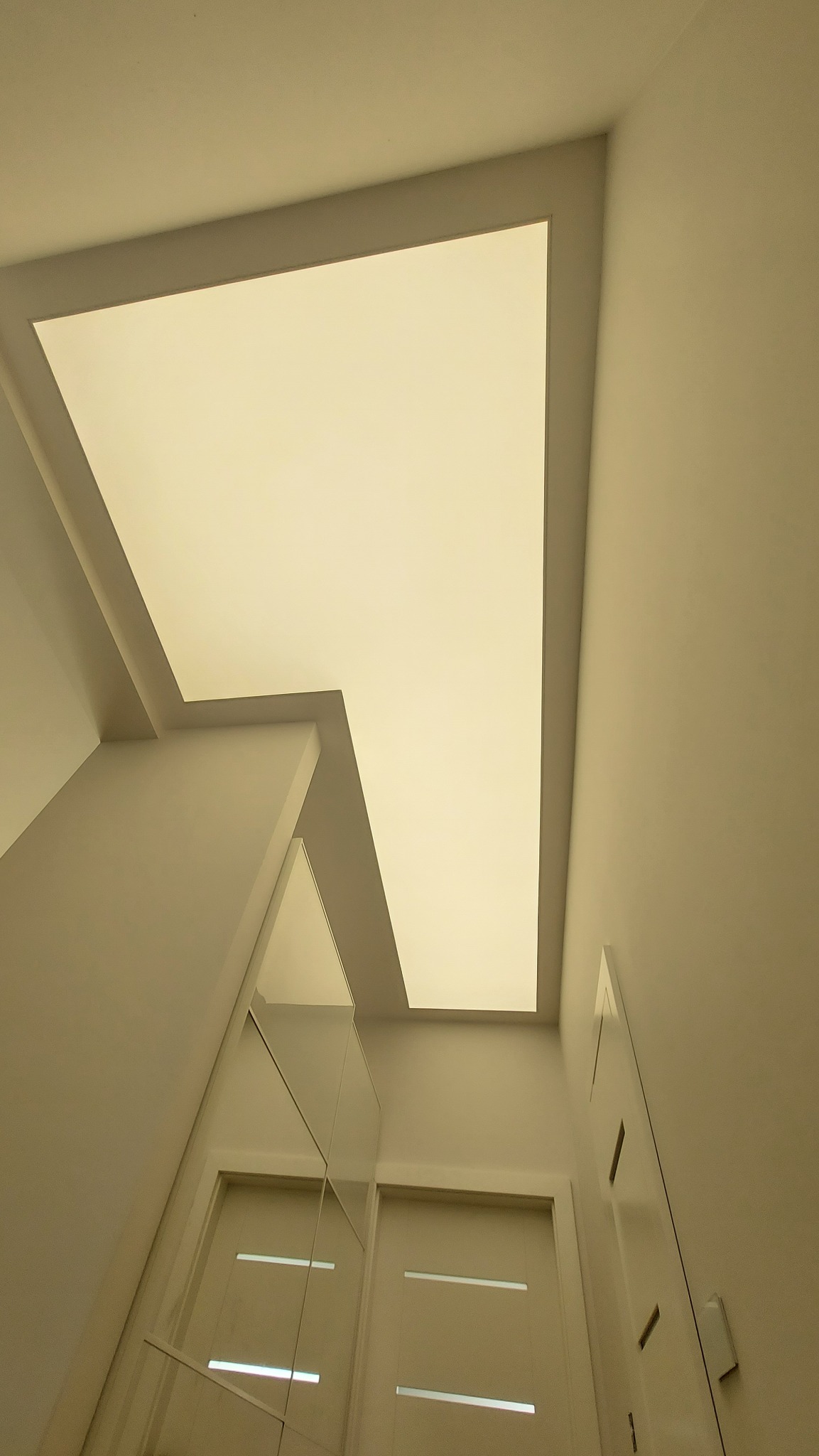Nowoczesny korytarz z podświetlanym sufitem o nieregularnym kształcie, widok z dołu, minimalistyczne drzwi w tle.