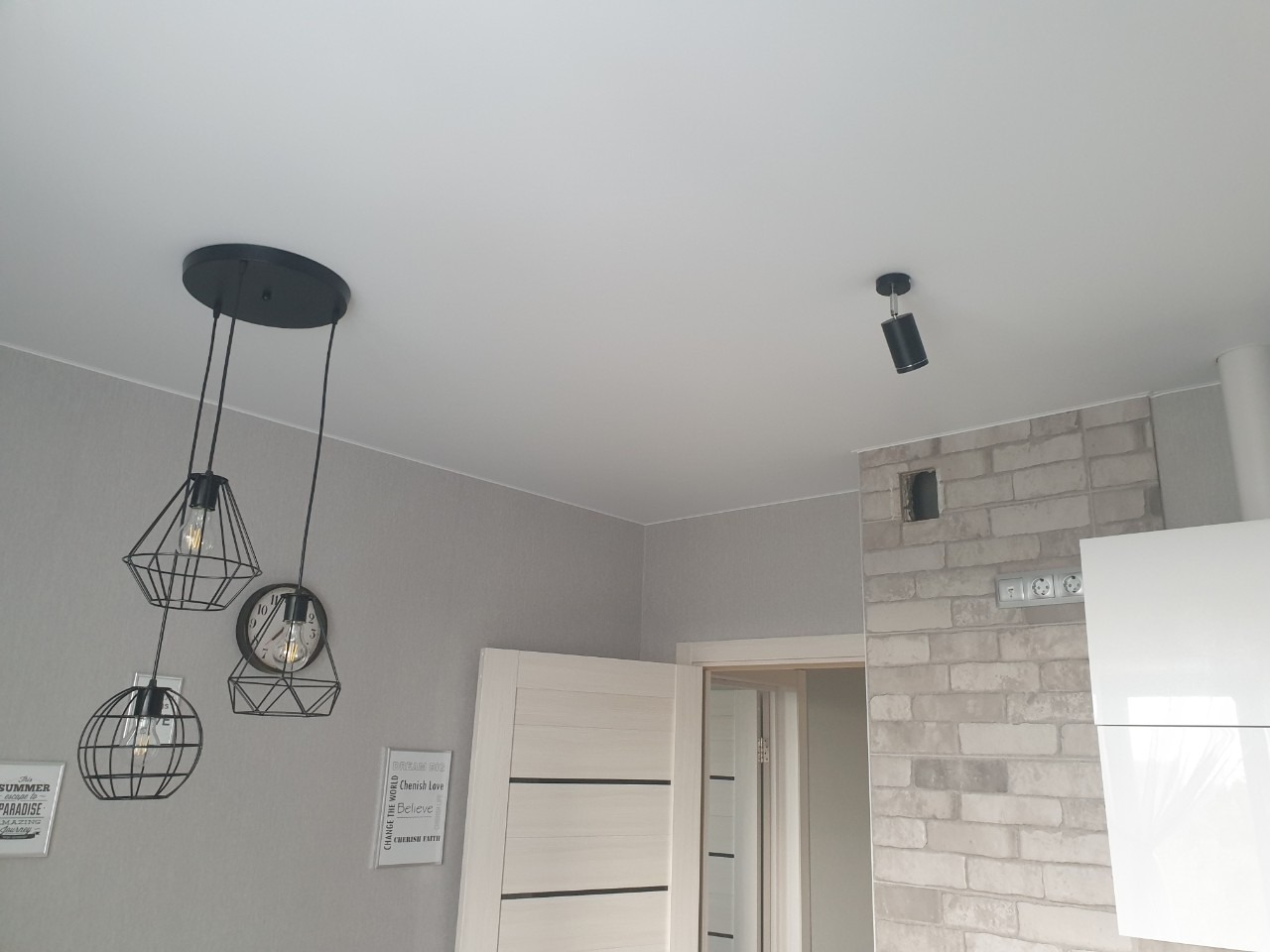 Nowoczesne oświetlenie sufitowe: czarna lampa wisząca z trzema kloszami w kształcie geometrycznych klatek i pojedynczy reflektor punktowy na białym suficie w szarym wnętrzu z dekoracyjną ścianą...