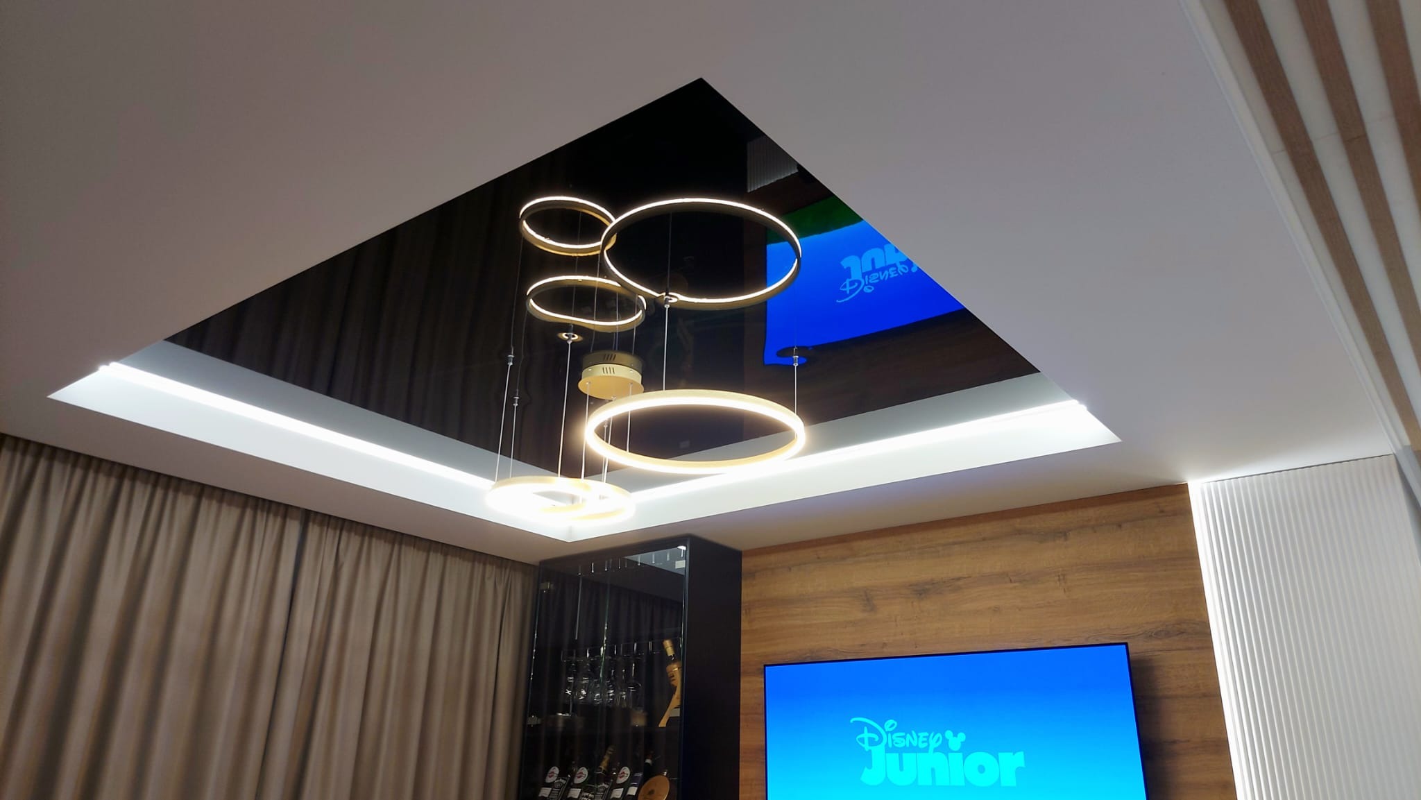 Nowoczesny salon z czarnym, błyszczącym sufitem, okrągłymi lampami wiszącymi i podświetleniem LED, odbijającym ekran telewizora.
