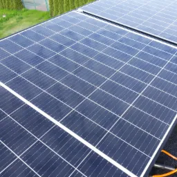 Czyste, lśniące panele słoneczne po umyciu, widoczne z góry, z fragmentem trawnika w tle i pomarańczowym przewodem u dołu kadru.