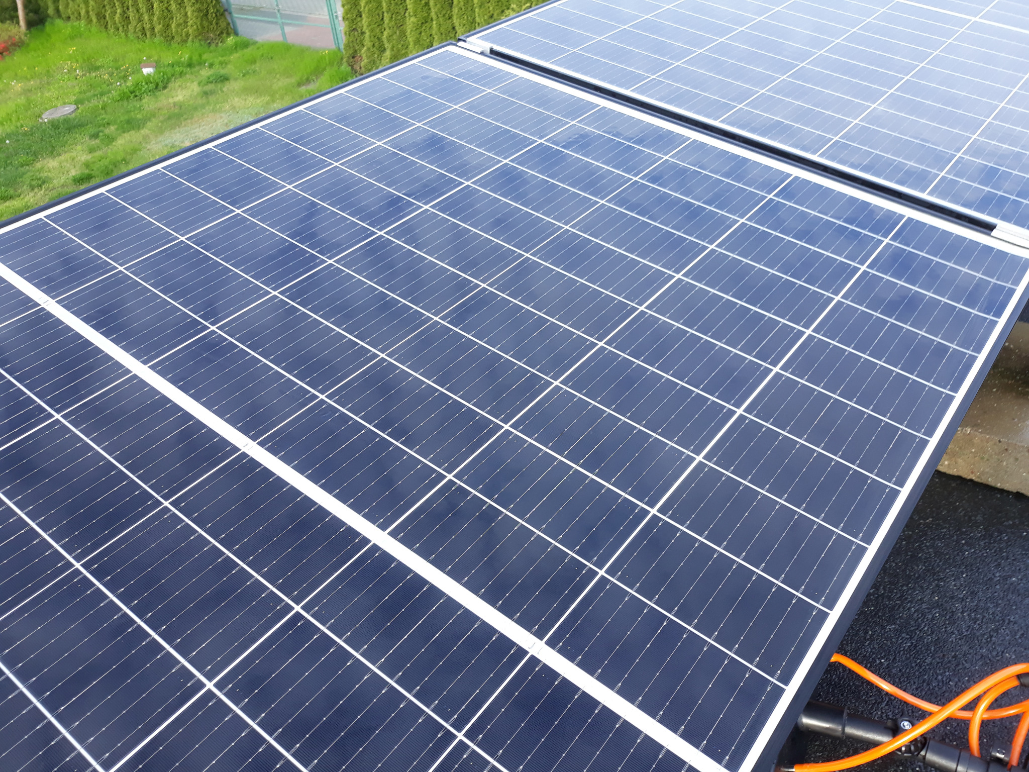 Czyste, lśniące panele słoneczne po umyciu, widoczne z góry, z fragmentem trawnika w tle i pomarańczowym przewodem u dołu kadru.