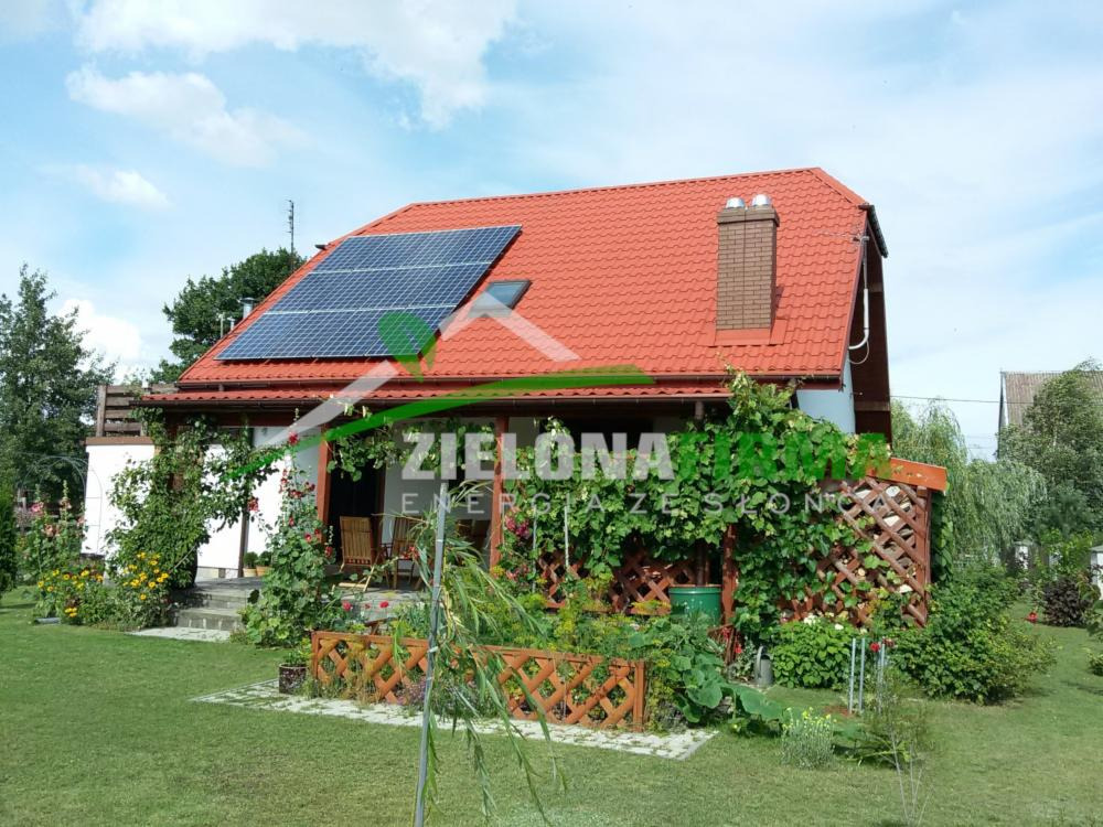 Dom jednorodzinny z czerwoną dachówką i panelami fotowoltaicznymi na dachu, otoczony bujną zielenią ogrodu, z widocznym logo firmy 'Zielona Energia' na elewacji.