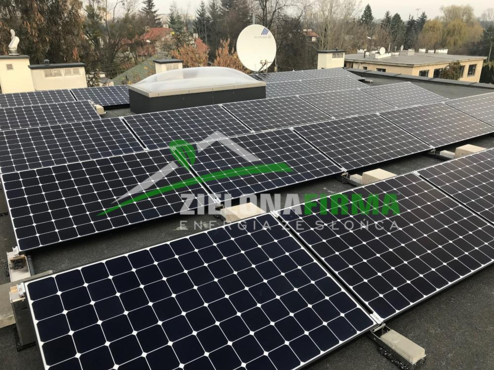 Instalacja paneli słonecznych na płaskim dachu budynku z widocznym świetlikiem i anteną satelitarną, tło stanowią drzewa i zabudowa mieszkalna.