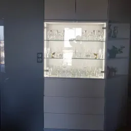 Biała, nowoczesna szafa z przeszkloną witryną, podświetlona od wewnątrz, eksponująca kolekcję szklanek i kieliszków o różnych kształtach i rozmiarach, z dodatkowymi półkami po prawej stronie.