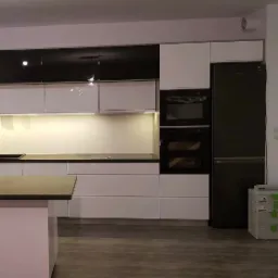 Nowoczesna kuchnia z białymi frontami dolnymi i czarnymi górnymi szafkami, oświetleniem LED pod szafkami, zabudowaną czarną lodówką i piekarnikiem, oraz szarym blatem.