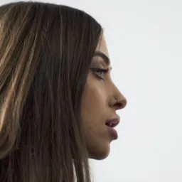 Profil kobiety z długimi, brązowymi włosami, widoczny kolczyk w nosie i delikatny makijaż oczu na jasnym tle.