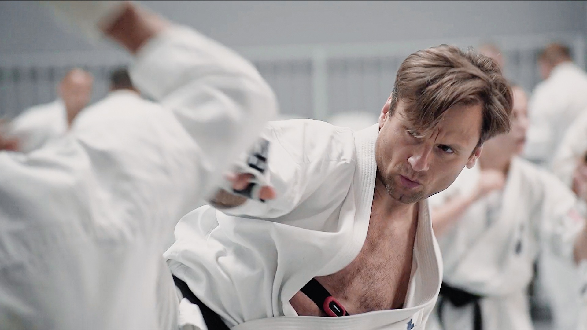 Mężczyzna w białym kimonie wykonuje dynamiczny ruch ręką, prawdopodobnie podczas treningu sztuk walki. W tle widoczni inni uczestnicy zajęć.