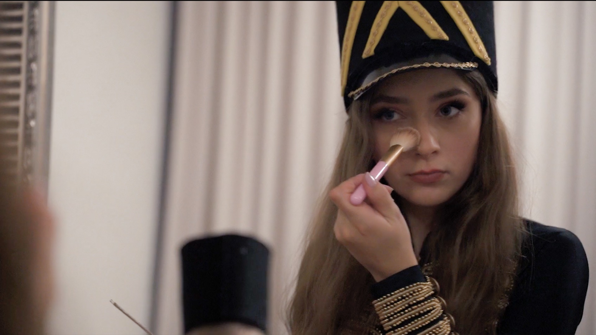 Młoda kobieta w kostiumie teatralnym, z czarną czapką ozdobioną złotymi detalami, nakłada puder pędzlem na nos, patrząc w lustro.