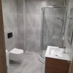 Nowoczesna łazienka po remoncie: szare płytki wielkoformatowe na ścianach i podłodze, kabina prysznicowa półokrągła ze srebrnymi profilami, wisząca toaleta z czarnym przyciskiem spłukującym, szafka...