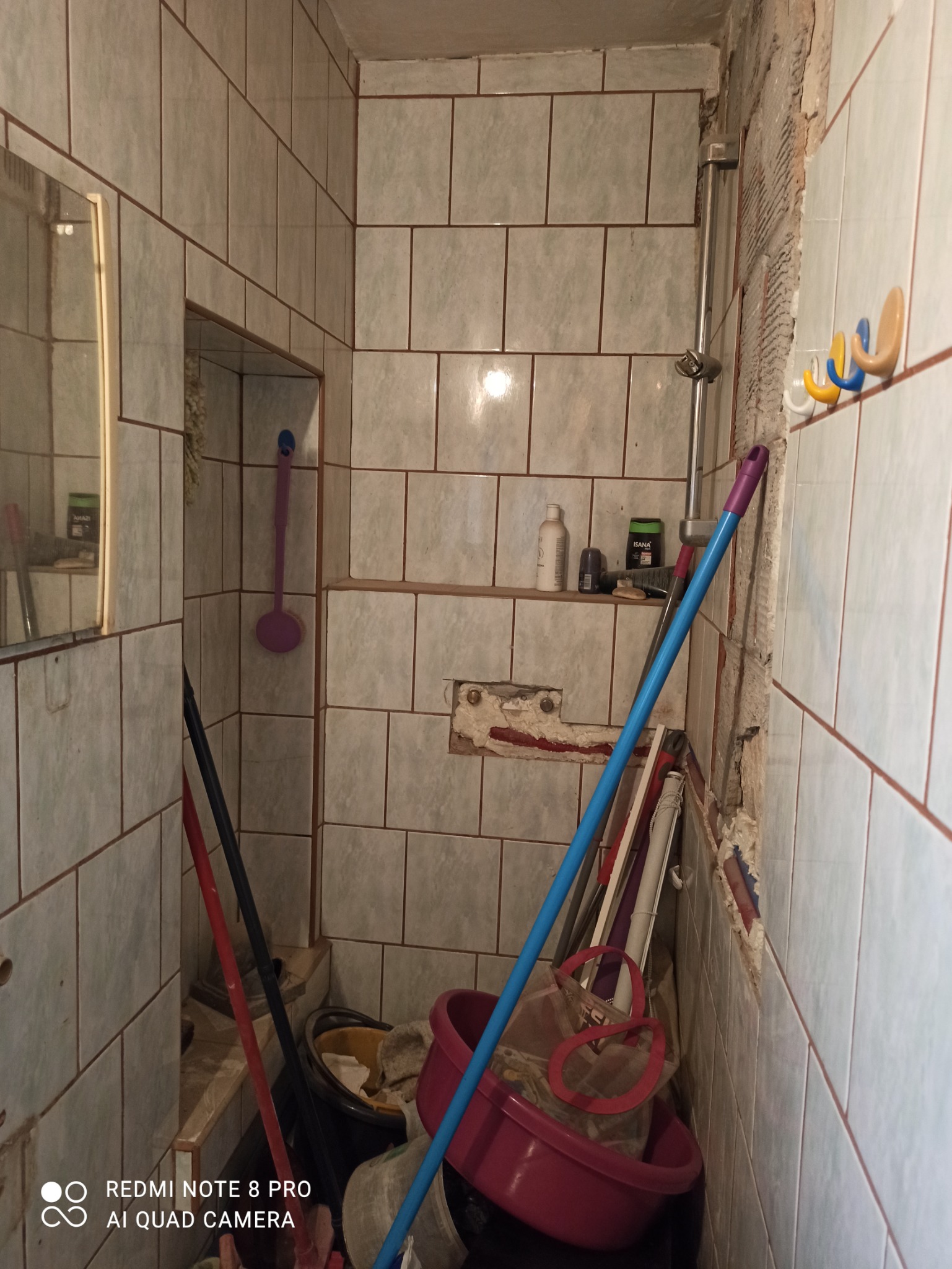 Łazienka w trakcie demontażu starych płytek; widoczne fragmenty ściany bez glazury, wiadro z odpadami, rurki, szczotka i akcesoria łazienkowe.