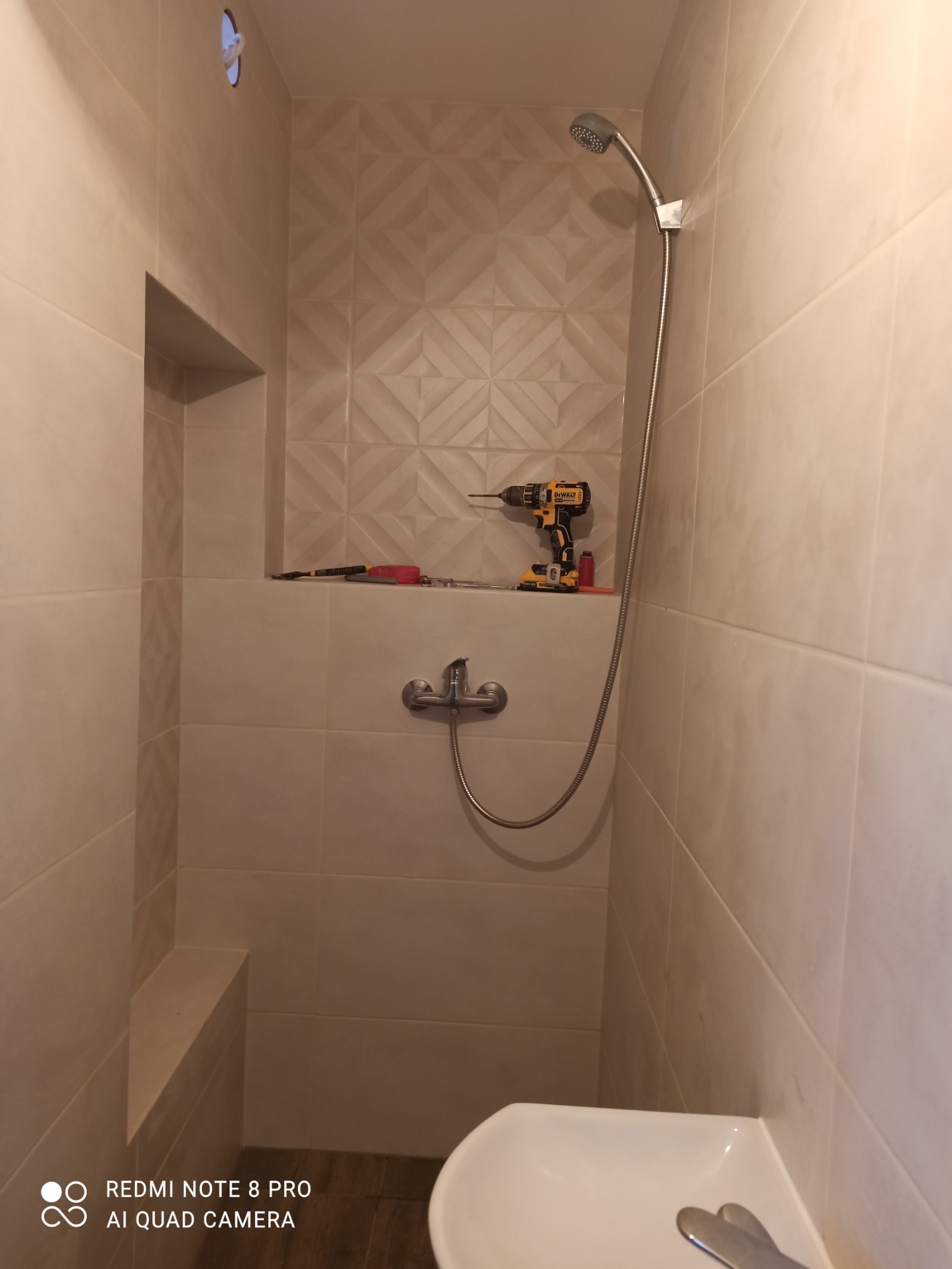 Wykończona kabina prysznicowa z beżowymi płytkami, widoczna bateria, słuchawka prysznicowa, półka w ścianie z narzędziami i wiertarką DeWalt, umywalka w dolnym rogu.