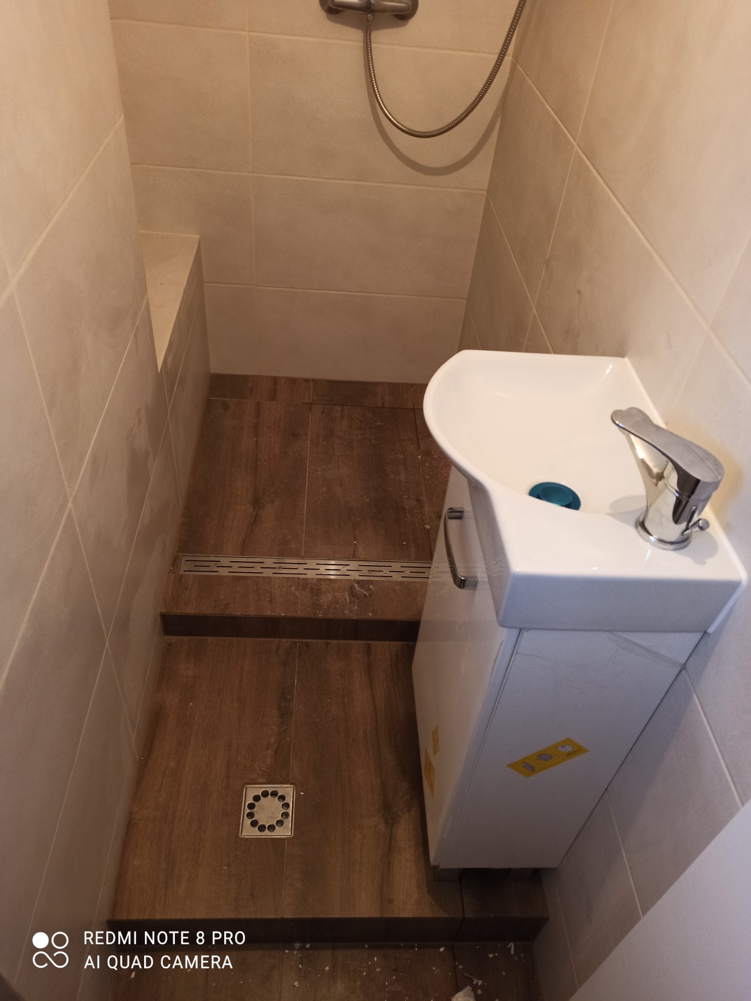 Wykończona łazienka z kabiną prysznicową typu walk-in, drewnopodobne płytki na podłodze, odpływ liniowy i punktowy, biała szafka z umywalką.