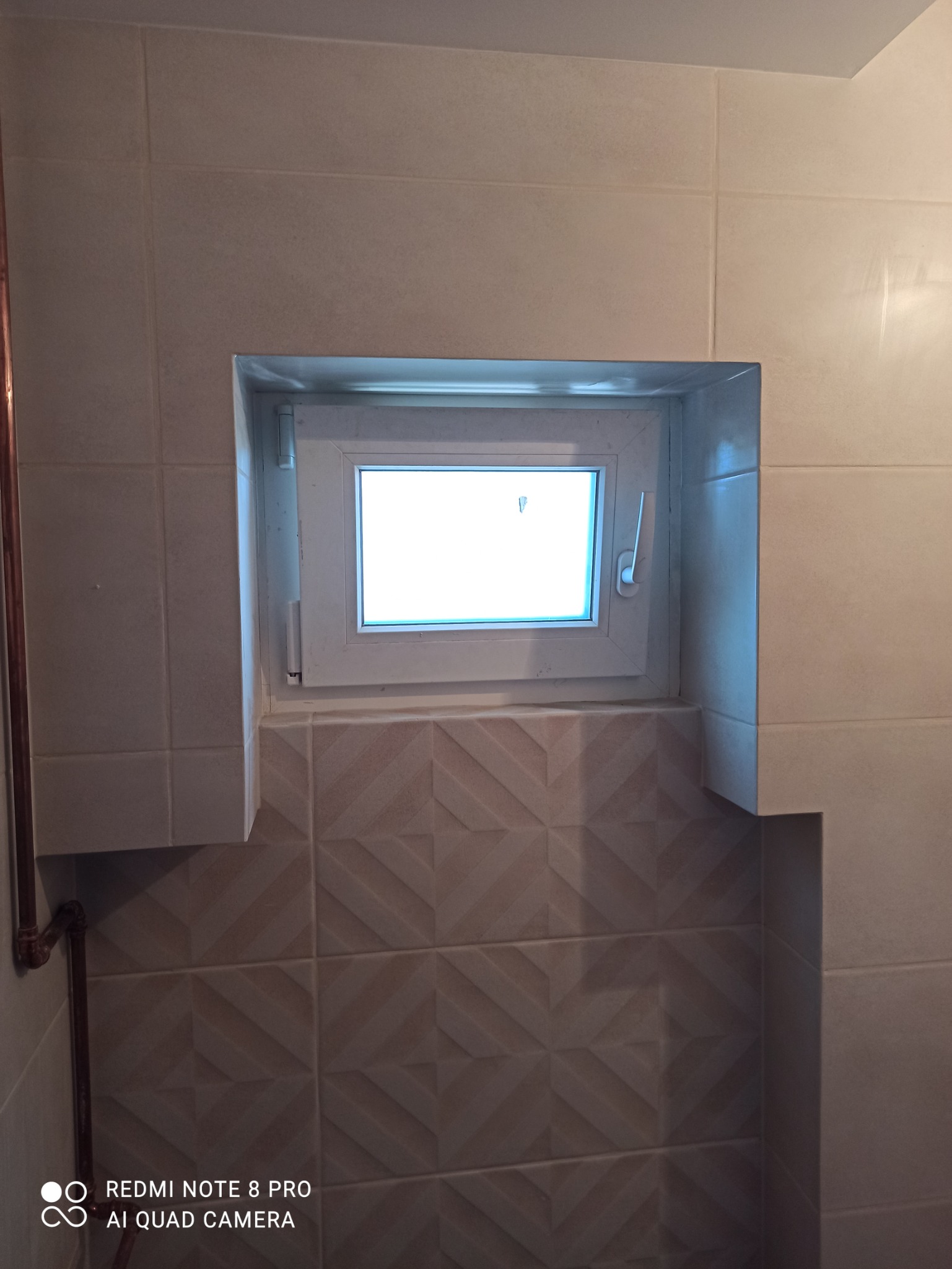 Wykończona wnęka okienna w łazience, obłożona jasnymi płytkami ceramicznymi z delikatnym wzorem, widoczna biała rama okienna i rury miedziane po lewej stronie.