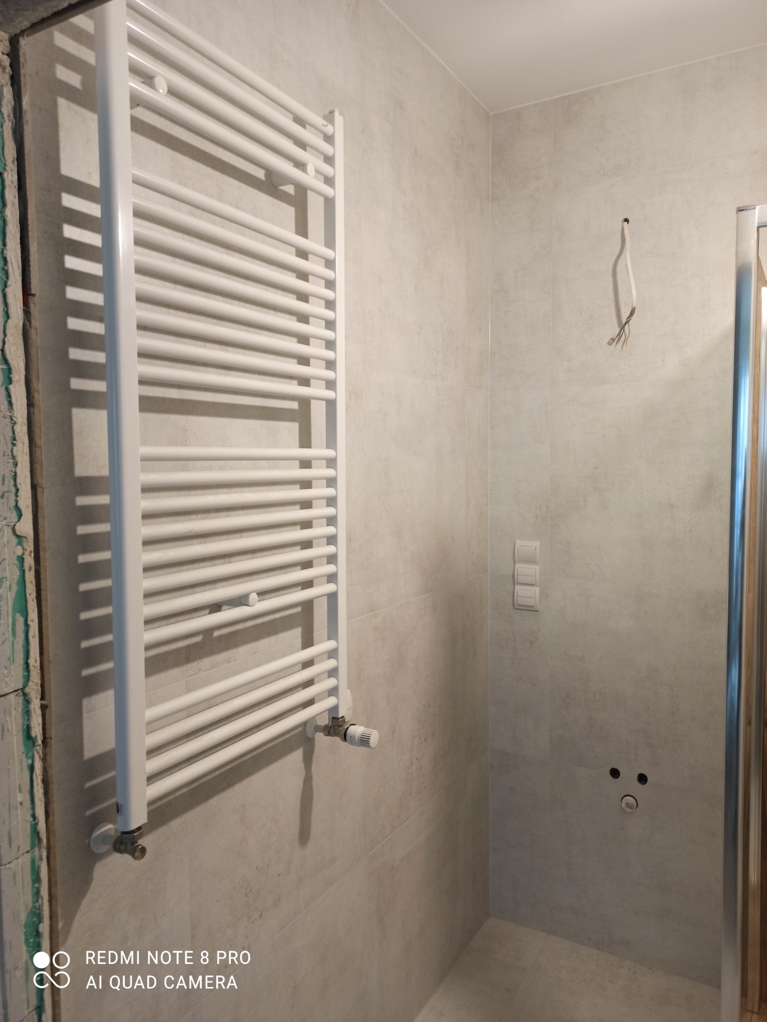 Biały, drabinkowy grzejnik łazienkowy zamontowany na szarej, kamiennej ścianie w trakcie prac wykończeniowych, widoczne niedokończone krawędzie ściany i wystające przewody.