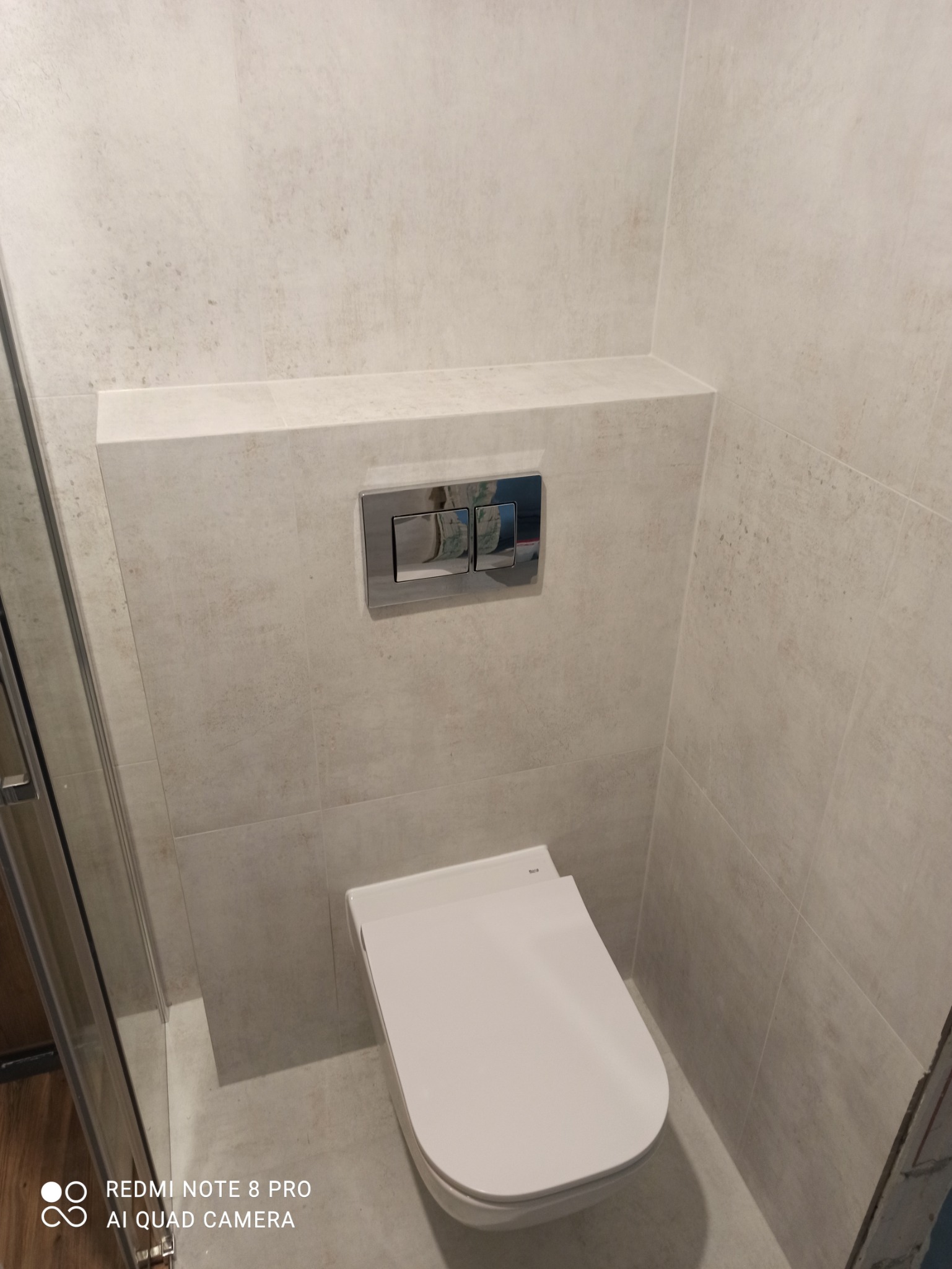 Nowoczesna toaleta podwieszana z chromowanym przyciskiem spłukującym w łazience wykończonej jasnymi, imitującymi kamień płytkami ceramicznymi.