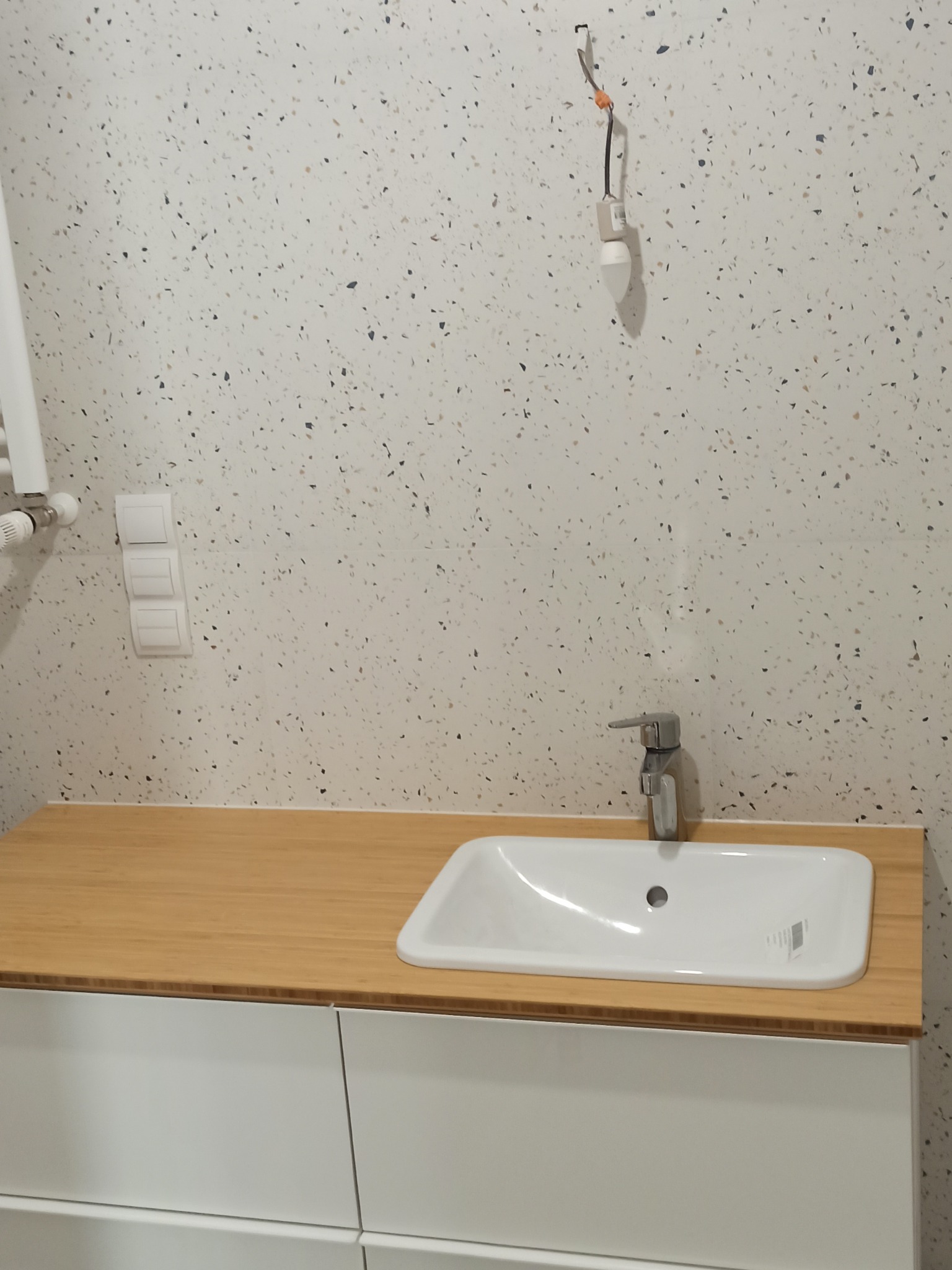 Łazienka w trakcie wykańczania: biała umywalka nablatowa z chromowaną baterią na blacie z jasnego drewna, białe szafki pod umywalką, ściana wyłożona płytkami imitującymi lastryko, widoczny włącznik...