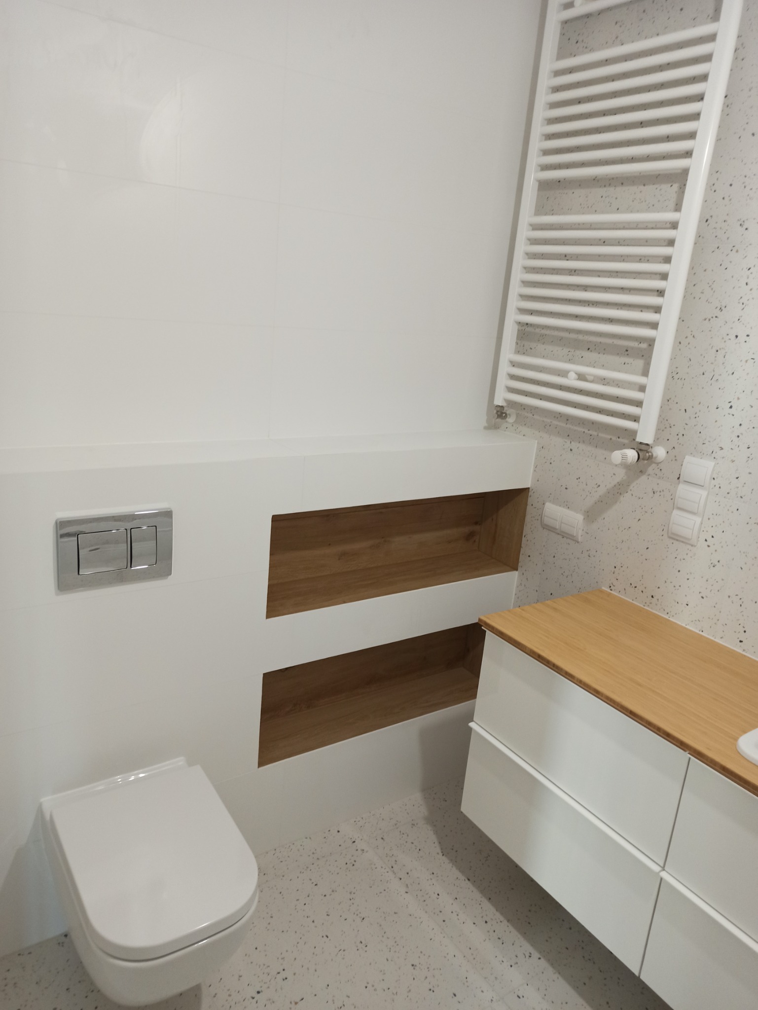 Nowoczesna łazienka z białymi płytkami, wiszącą toaletą z chromowanym przyciskiem, zabudowanymi półkami z drewnianym wykończeniem, białą szafką pod umywalkę z drewnianym blatem i grzejnikiem...