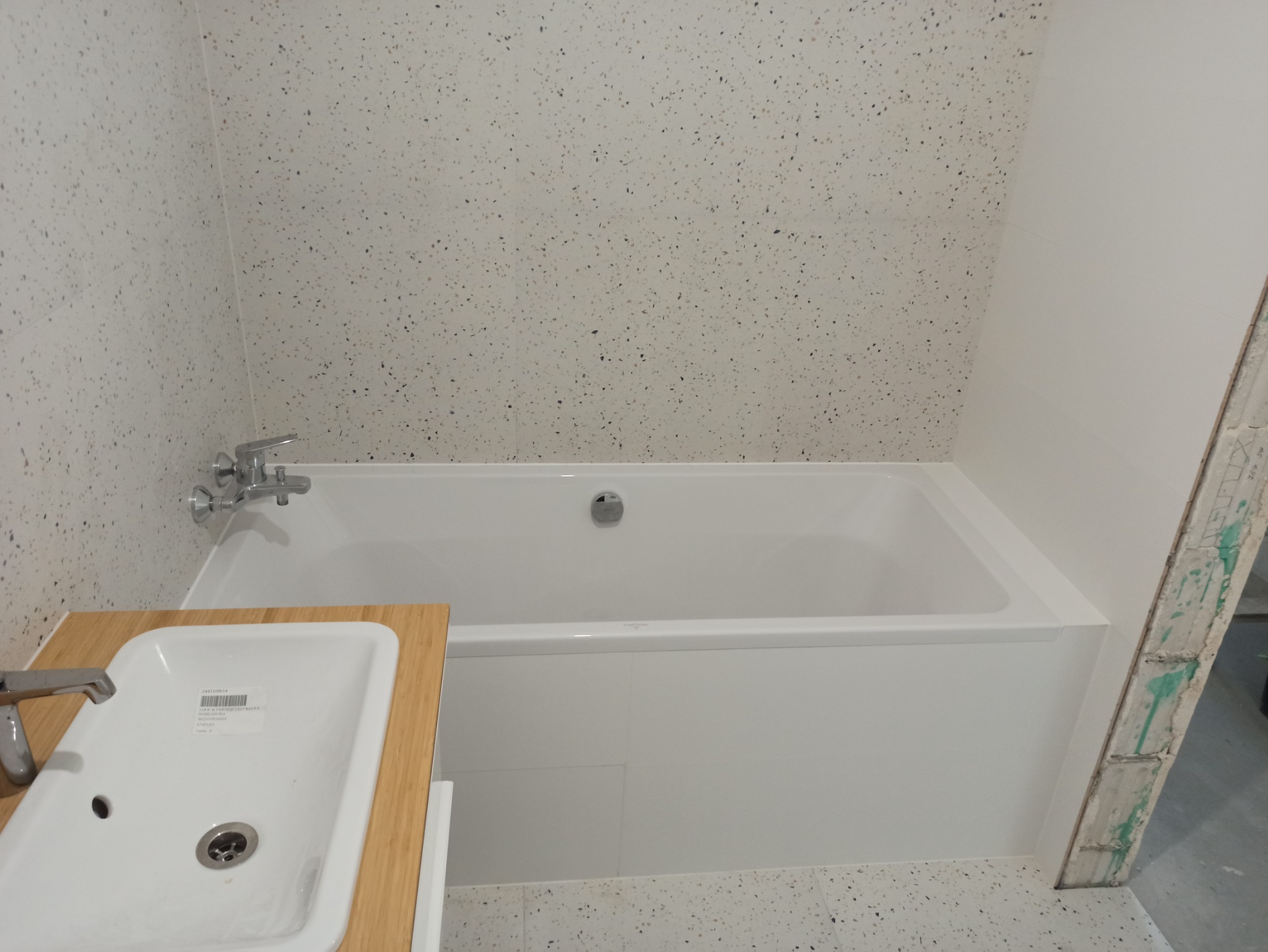 Nowa, biała wanna z chromowaną baterią i odpływem, umywalka z blatem z jasnego drewna, ściany i podłoga wyłożone jasnymi płytkami z ciemnymi plamkami, widoczny fragment ściany w stanie surowym.