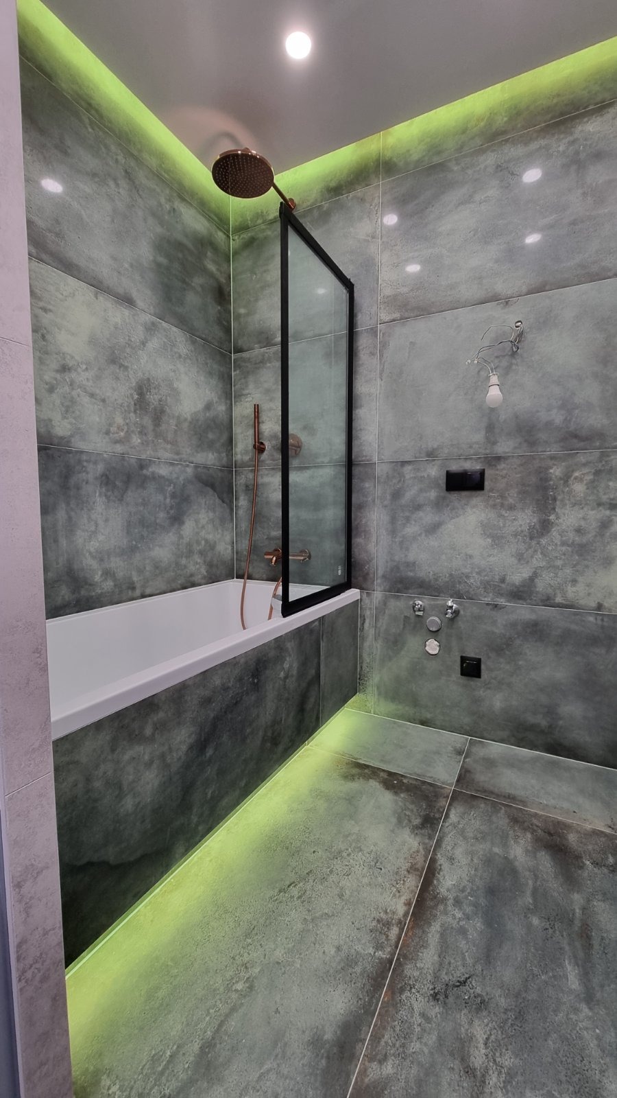 Nowoczesna łazienka z szarymi płytkami imitującymi beton, podświetleniem LED, kabiną prysznicową z czarną ramą i armaturą w kolorze miedzi.