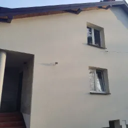 Jasna elewacja domu jednorodzinnego z brązowym dachem, widoczne okna z białymi ramami i kolumny wejściowe z elementami dekoracyjnymi imitującymi kamień.