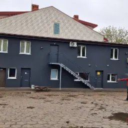 Dwupiętrowy budynek pomalowany na ciemny grafit z białymi ramami okiennymi i zewnętrznymi klimatyzatorami, widoczne dwie osoby w odzieży roboczej na brukowanym dziedzińcu, białe auto zaparkowane...