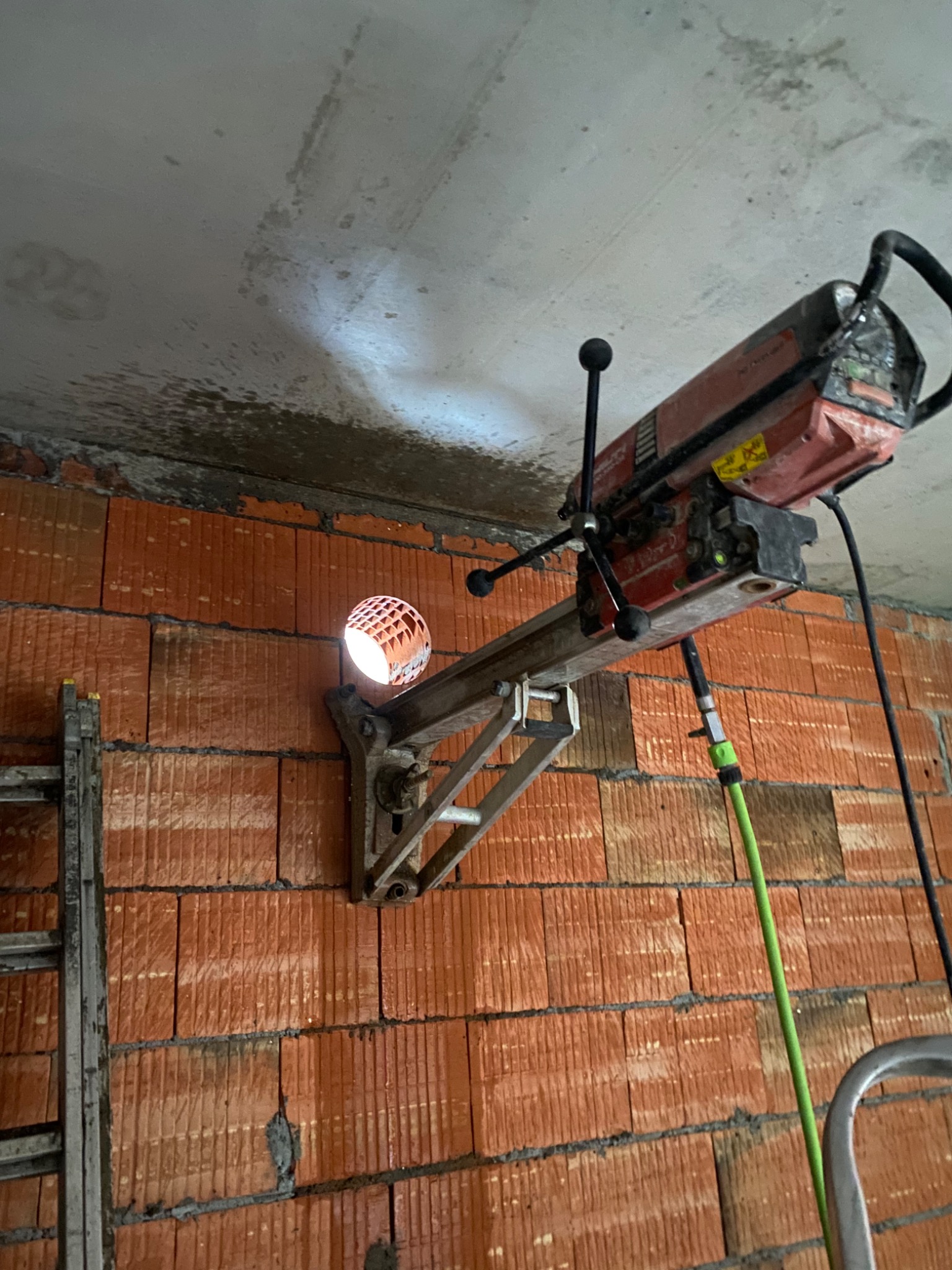 Wiercenie rdzeniowe w czerwonej ceglanej ścianie z użyciem wiertnicy z mocowaniem, widoczny otwór z kratownicą w środku, przewód wodny chłodzący i drabina oparta o ścianę.