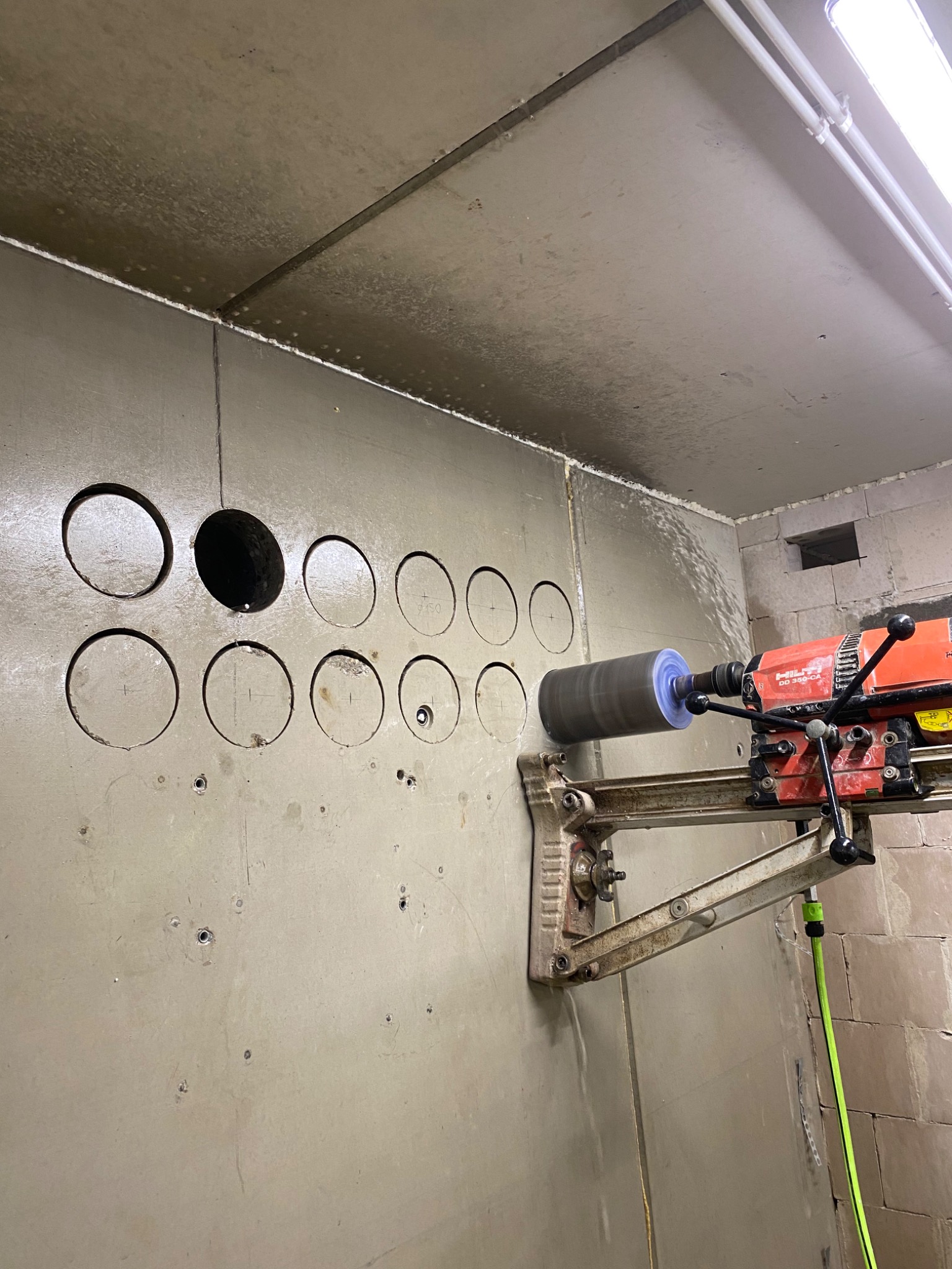 Wiertnica diamentowa Hilti DD 350-CA w trakcie wykonywania otworów w szarej ścianie, widoczne liczne wywiercone otwory o różnych średnicach, ślady wody po chłodzeniu wiertła.