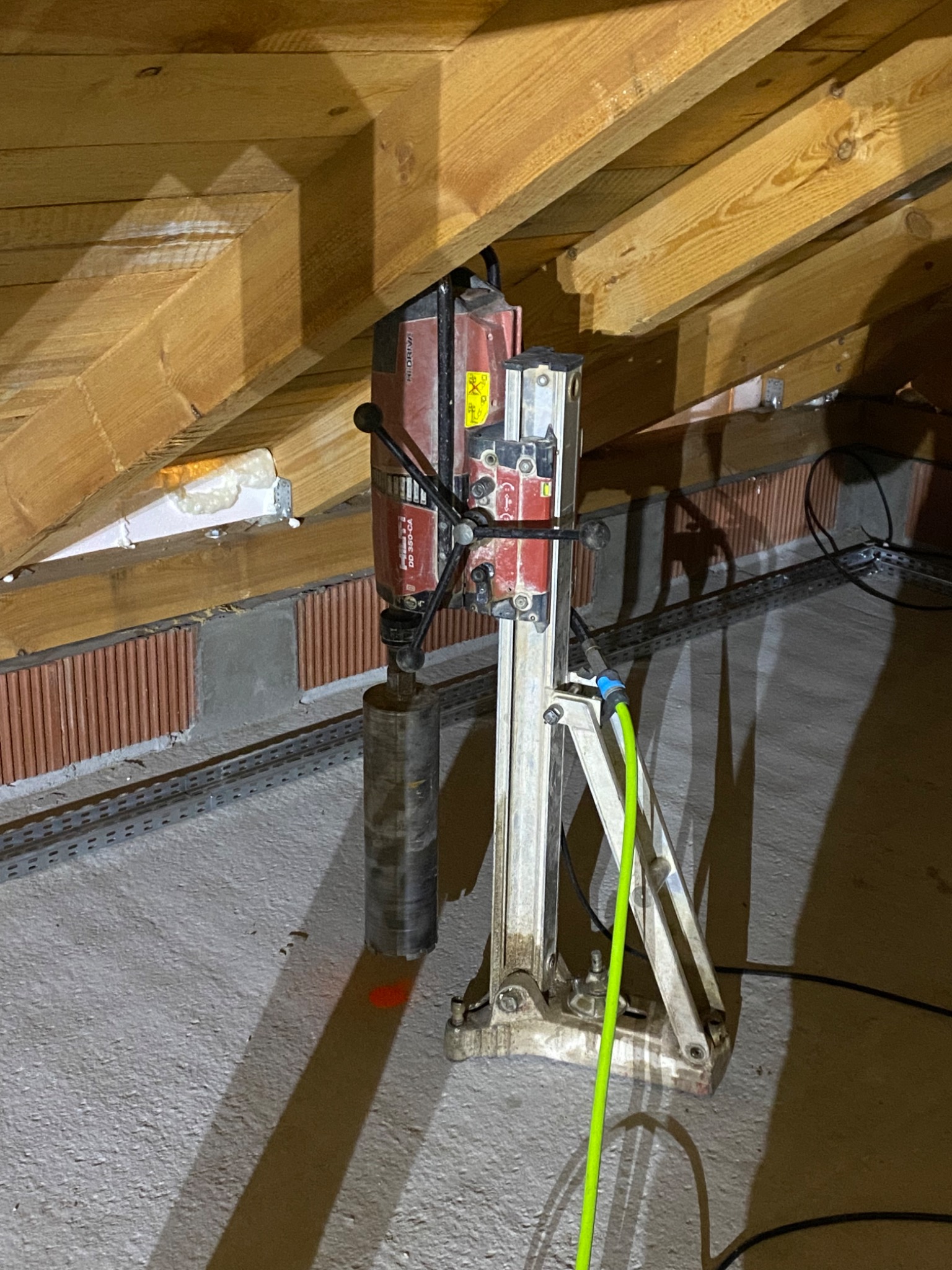 Wiertnica diamentowa na statywie, przygotowana do wiercenia rdzeniowego w betonowej posadzce, widoczne drewniane belki stropowe i fragment ściany z cegieł.
