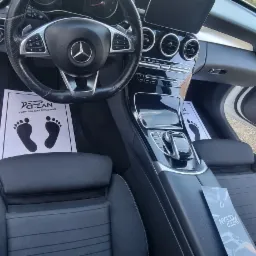Wnętrze białego samochodu Mercedes-Benz po czyszczeniu, z papierowymi matami podłogowymi z nadrukowanymi stopami i logo firmy czyszczącej Deep Clean, widoczne fotele i deska rozdzielcza.
