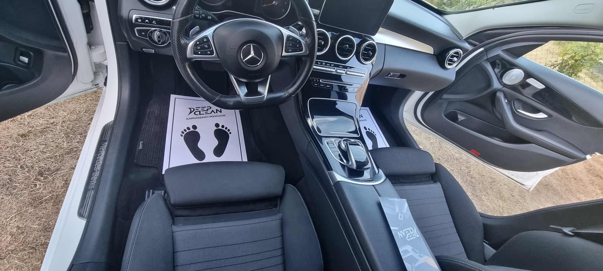 Wnętrze białego samochodu Mercedes-Benz po czyszczeniu, z papierowymi matami podłogowymi z nadrukowanymi stopami i logo firmy czyszczącej Deep Clean, widoczne fotele i deska rozdzielcza.