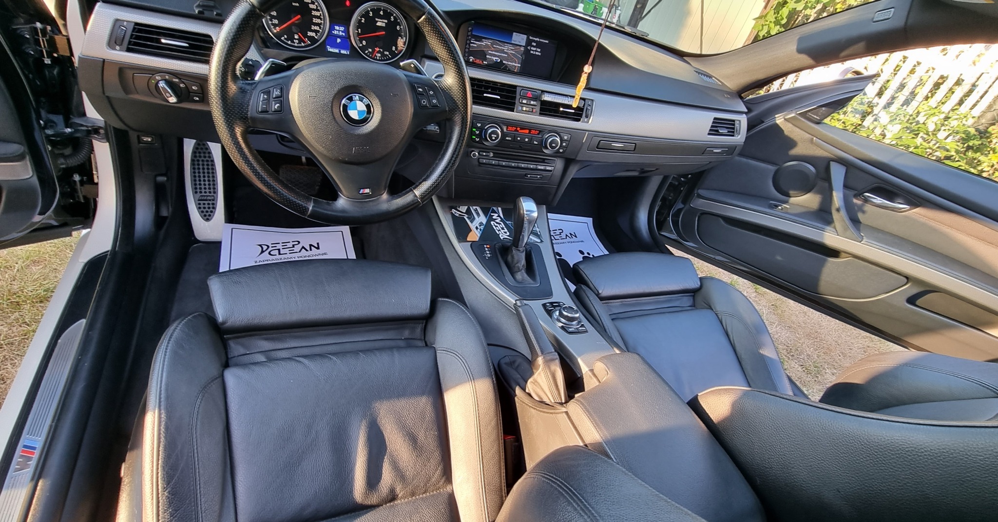 Wnętrze czarnego BMW po detailingu, widoczne skórzane fotele, deska rozdzielcza i kierownica, z ulotkami firmy detailingowej na siedzeniach.