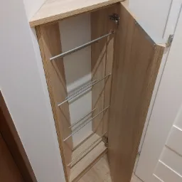 Narożna szafka z otwartymi drzwiami, wykonana z drewna w jasnym odcieniu dębu, z metalowymi relingami wewnątrz, zamontowana przy białej ścianie i białych drzwiach.