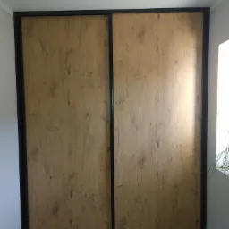 Dwudrzwiowa szafa wnękowa z przesuwnymi drzwiami o fakturze jasnego drewna, otoczona czarną ramą, oświetlona punktowym światłem sufitowym.